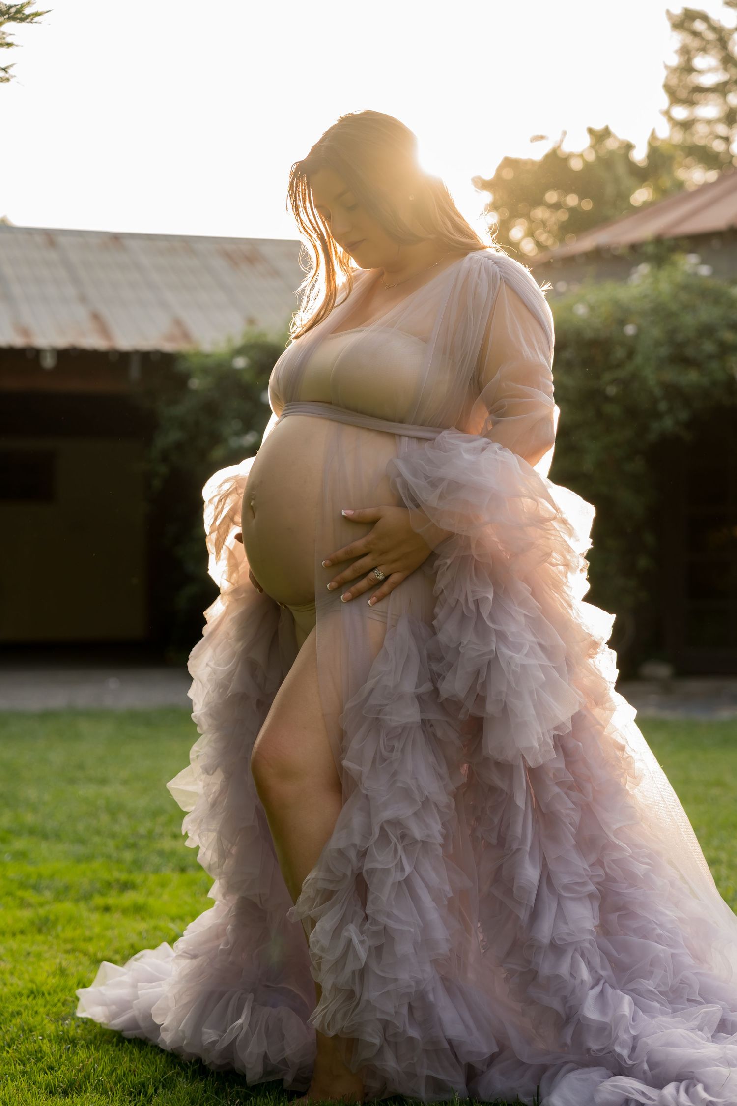 Expectant mother in flowing lavender tulle gown poses in golden sunlight on grassy lawn at sunset.