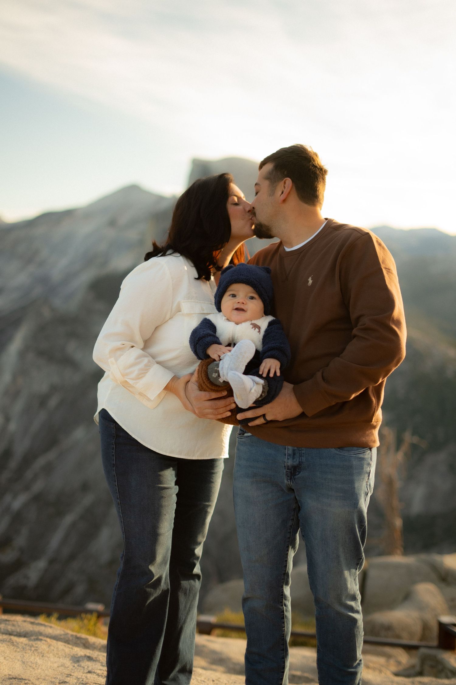 Family embraces while holding baby in cozy winter outfits against mountainous backdrop.