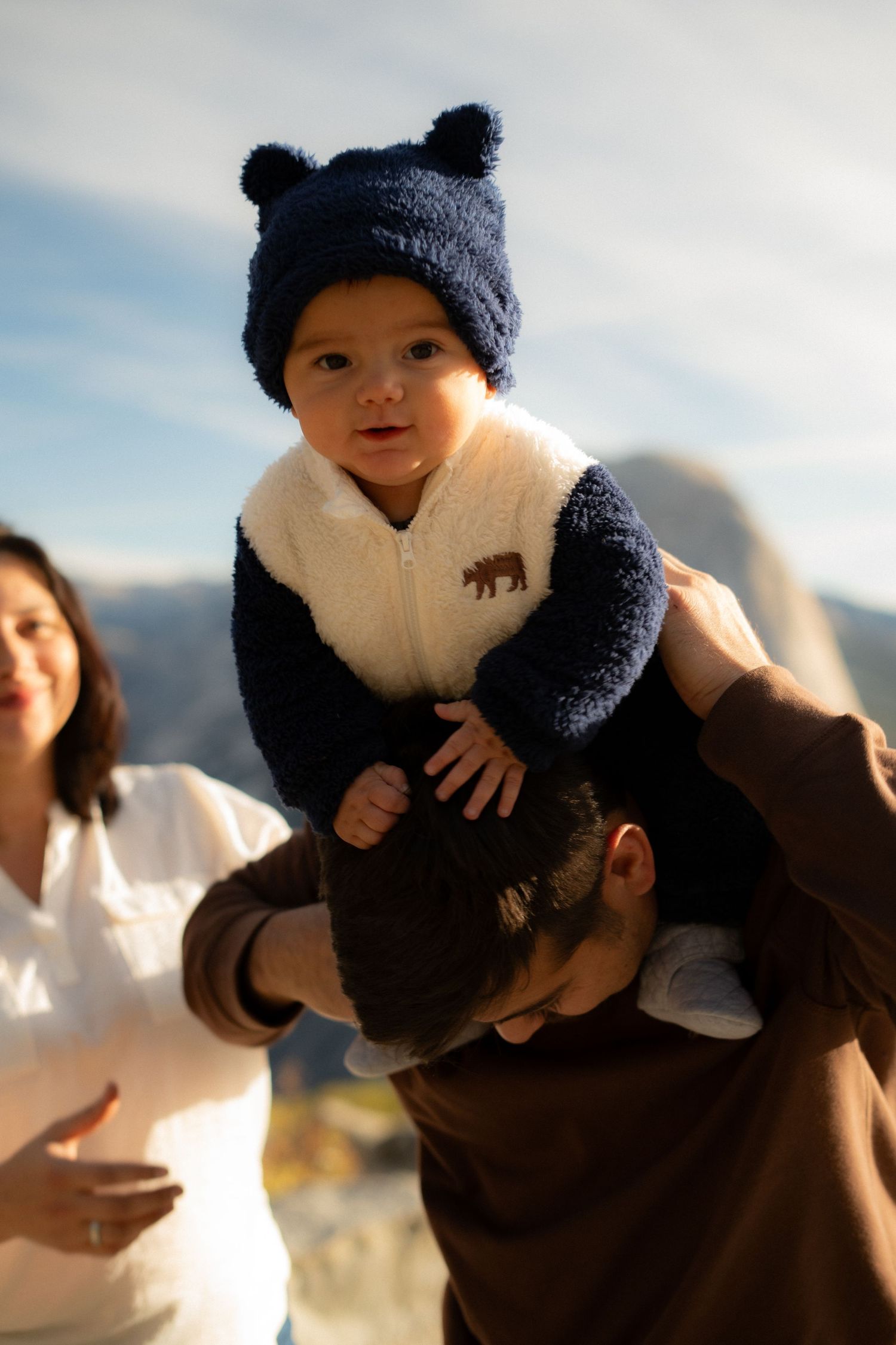 Baby in winter clothing smiles while being held up against mountain backdrop.