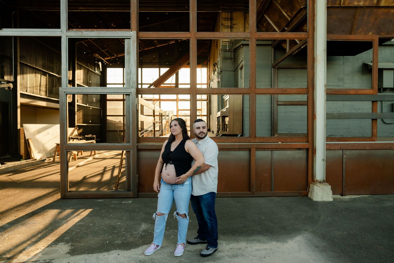 A couple stands together in an industrial setting with rustic metal structures and warm sunlight casting long shadows.