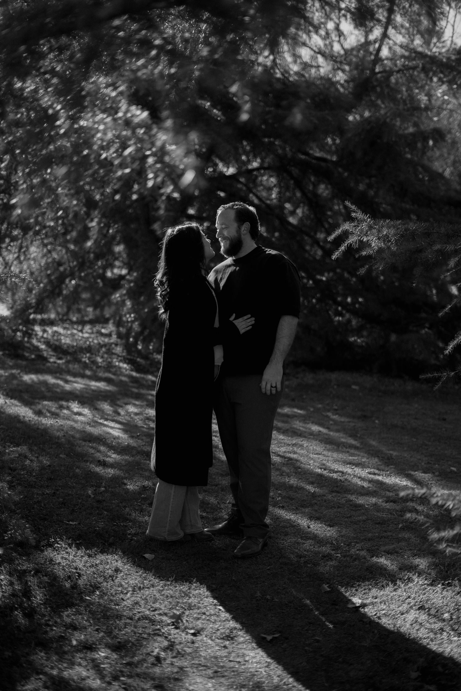A black and white photo of a couple standing close together in a wooded setting.