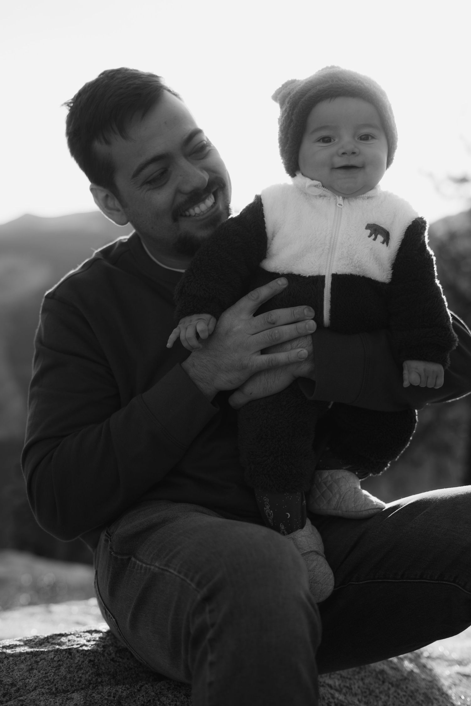 Baby in winter clothing smiling while being held outdoors against mountain backdrop.