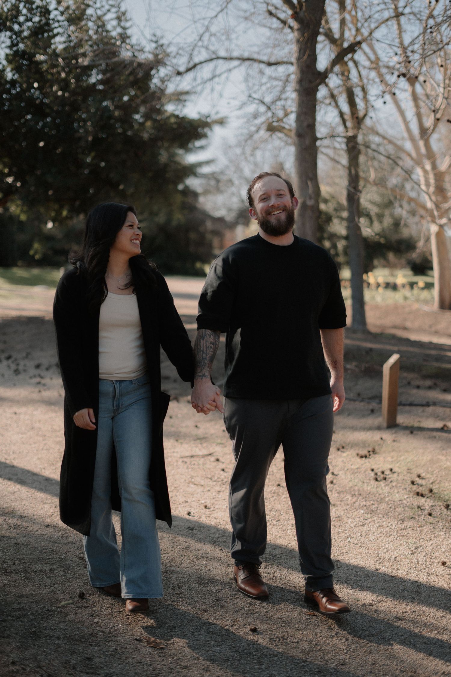 A couple walking hand in hand along a sunny park path in casual attire.