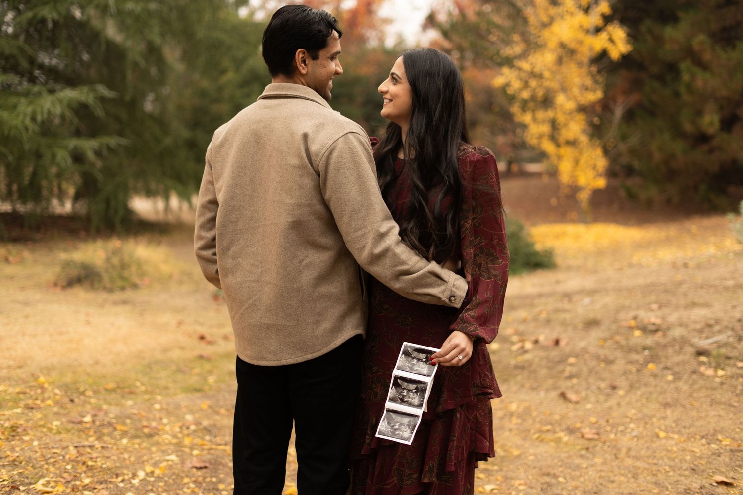 A couple holds sonogram photos while standing together in an autumn setting with fallen leaves.