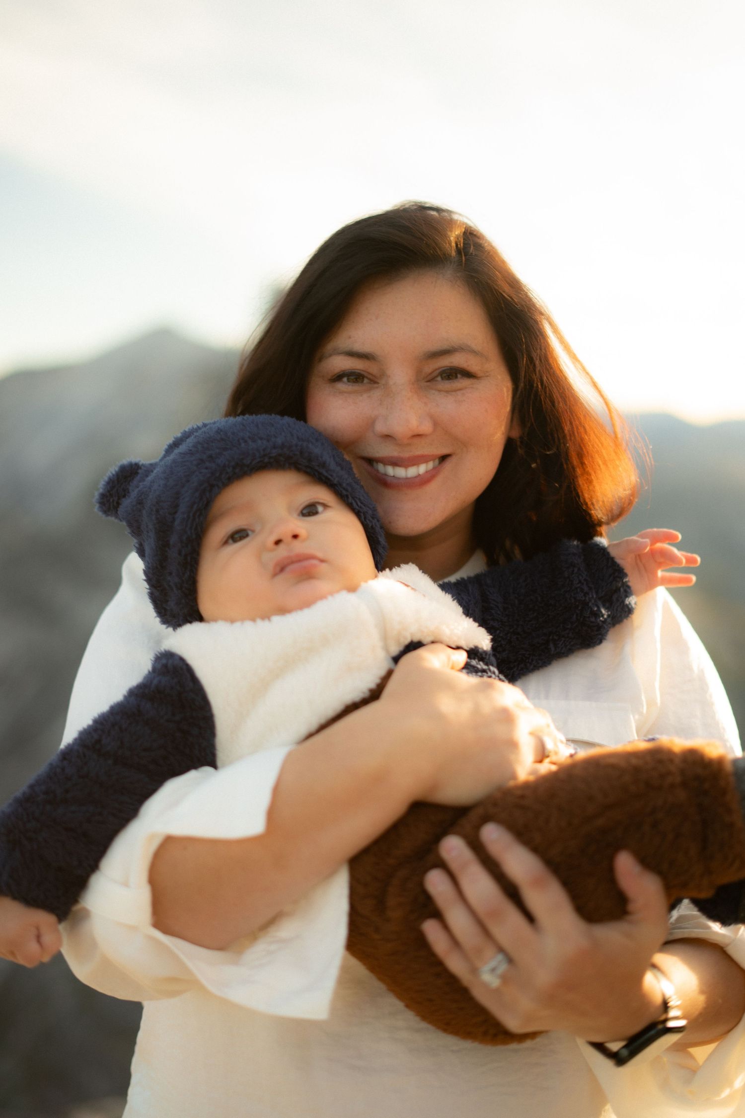 Mother and baby in mountain setting share joyful moment while holding stuffed toy.