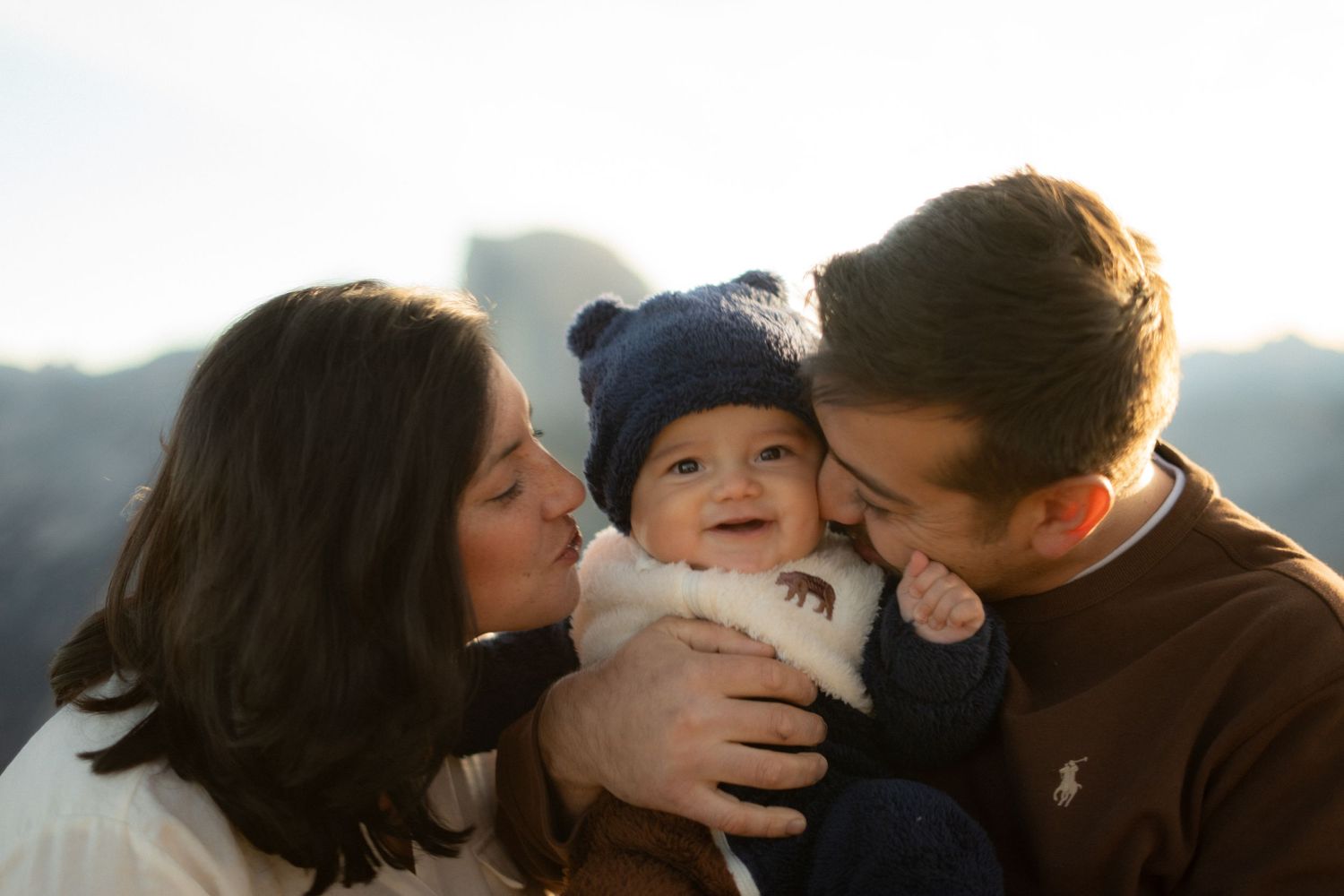 A couple shares a tender moment with their baby outdoors against a mountain backdrop.