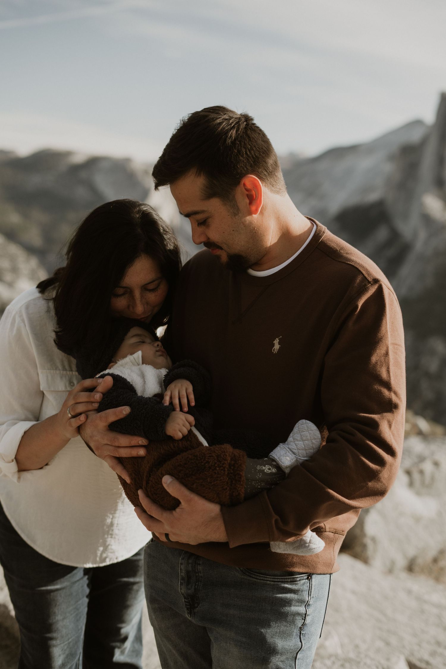 Candid family moment captured against mountain scenery as parents gaze at their infant.
