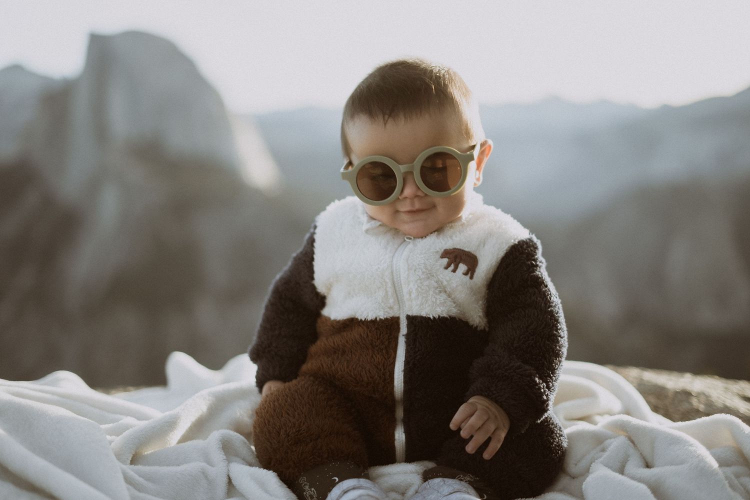 Baby wearing sunglasses and fuzzy outfit sits on white blanket with mountains behind.