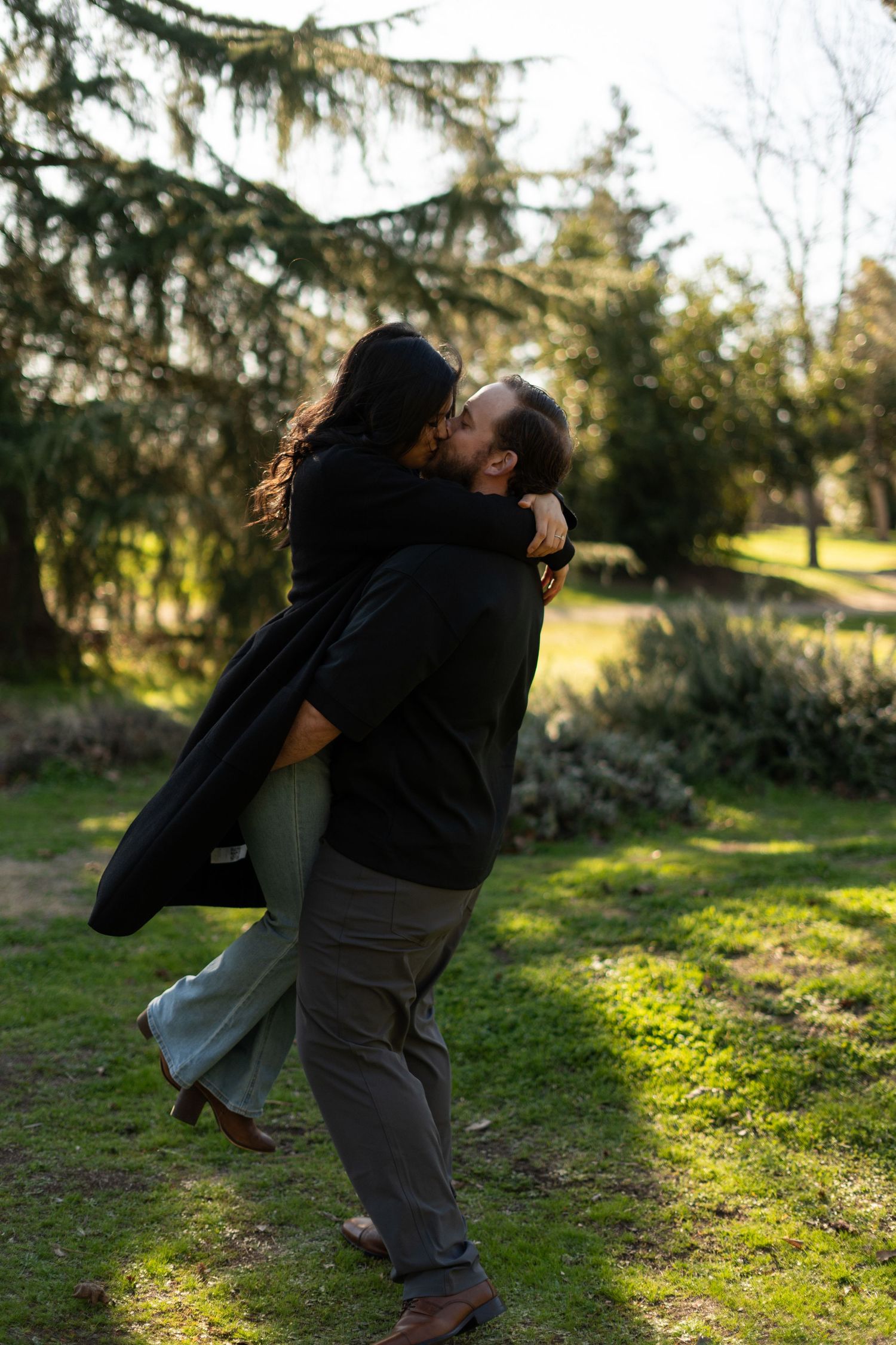 A romantic couple embracing in a sunlit garden with evergreen trees in the background.