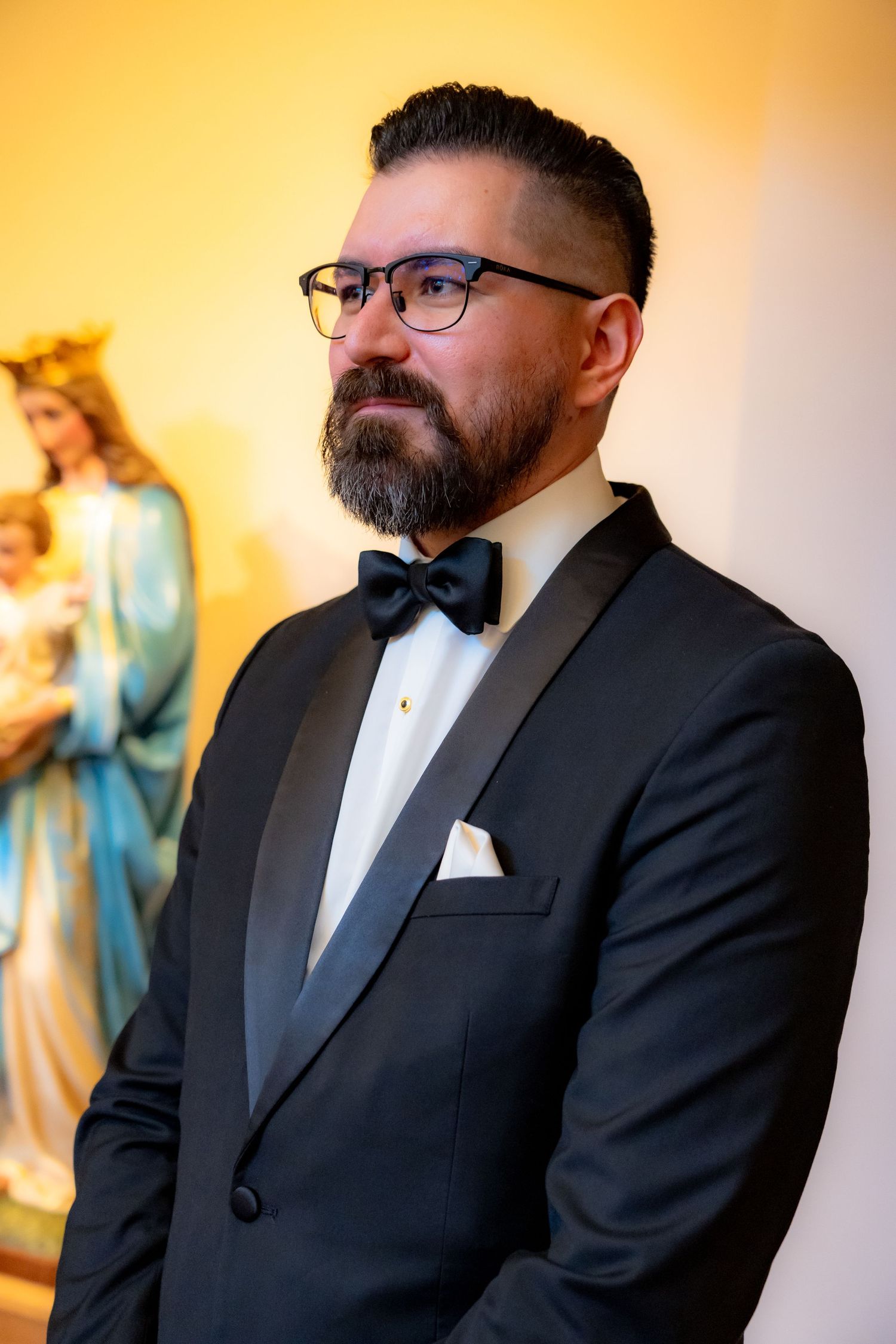 Groom in classic black tuxedo stands beside religious statue with warm lighting.