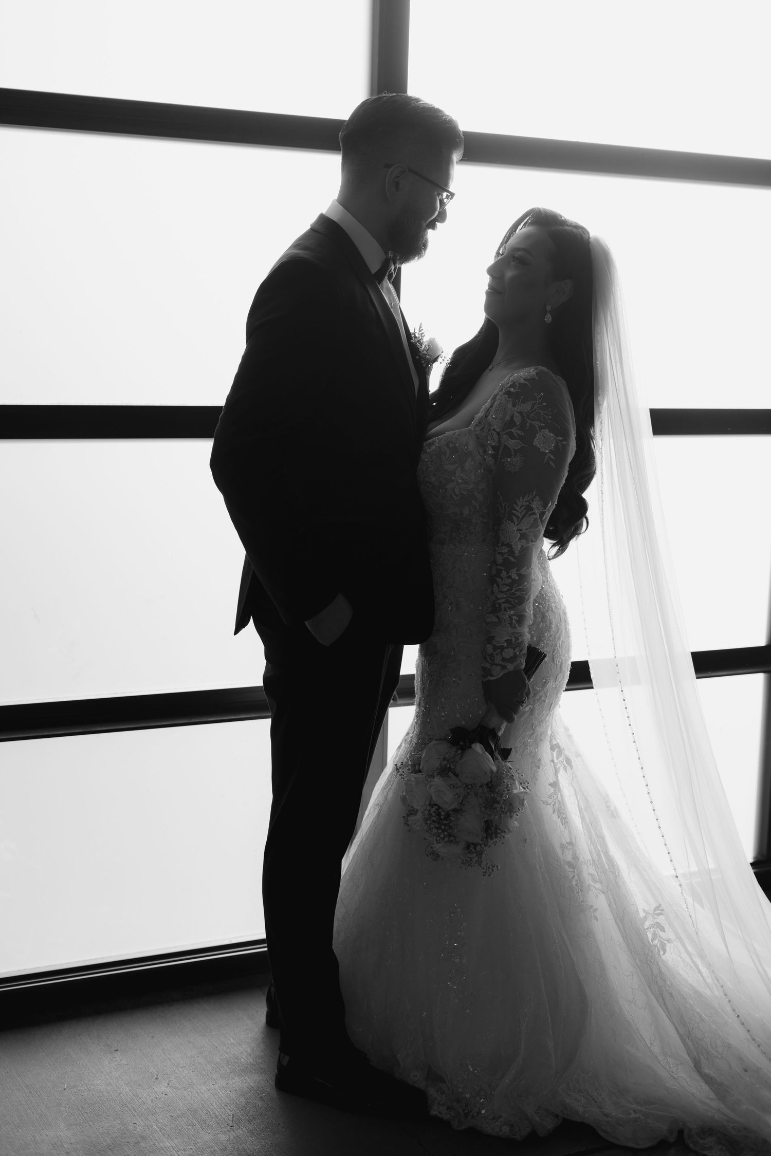 Black and white silhouettes of a couple in wedding attire standing by a window.
