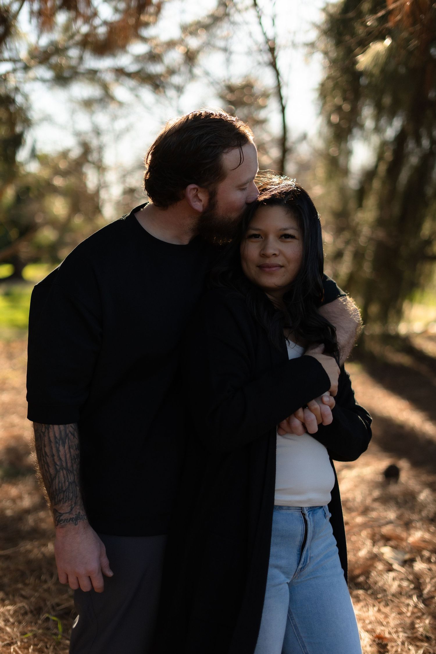 A couple dressed in casual wear embraces in a sunlit forest setting.