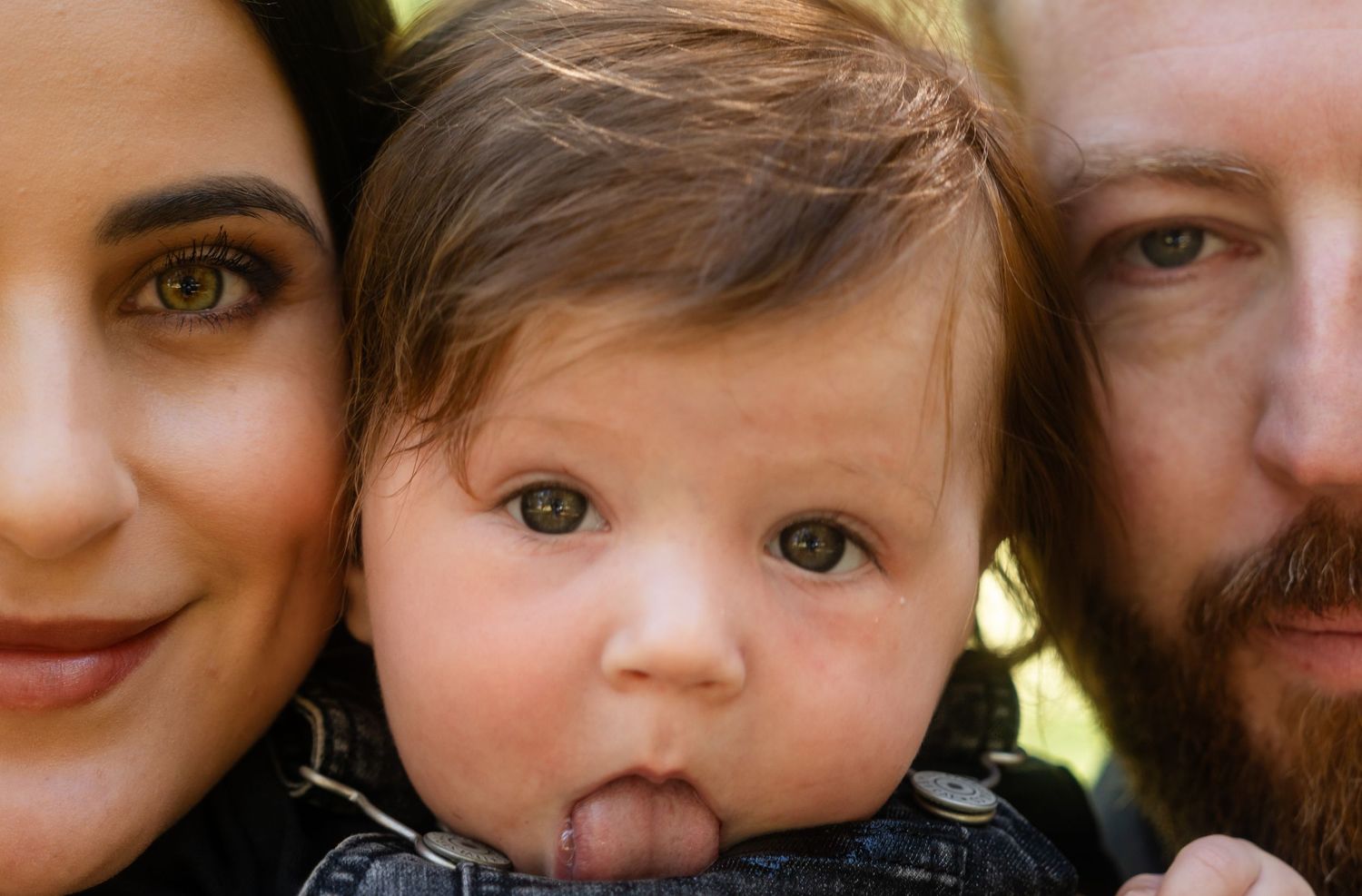 A happy family portrait featuring a dark-haired woman, a bearded man, and a cute baby sticking out their tongue.