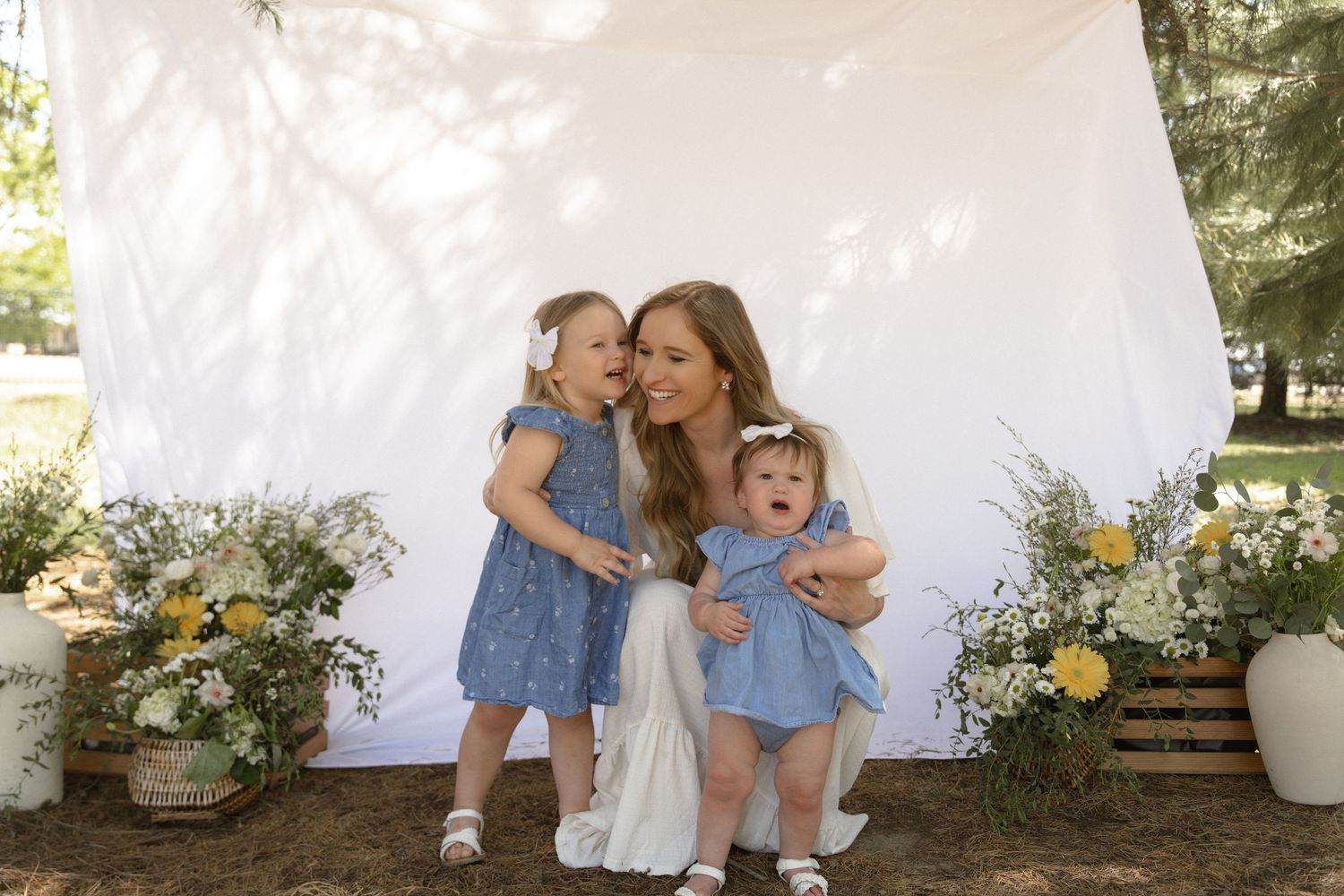 A smiling woman in white kneels outdoors with two toddler girls in blue dresses, surrounded by yellow flowers and white backdrop.