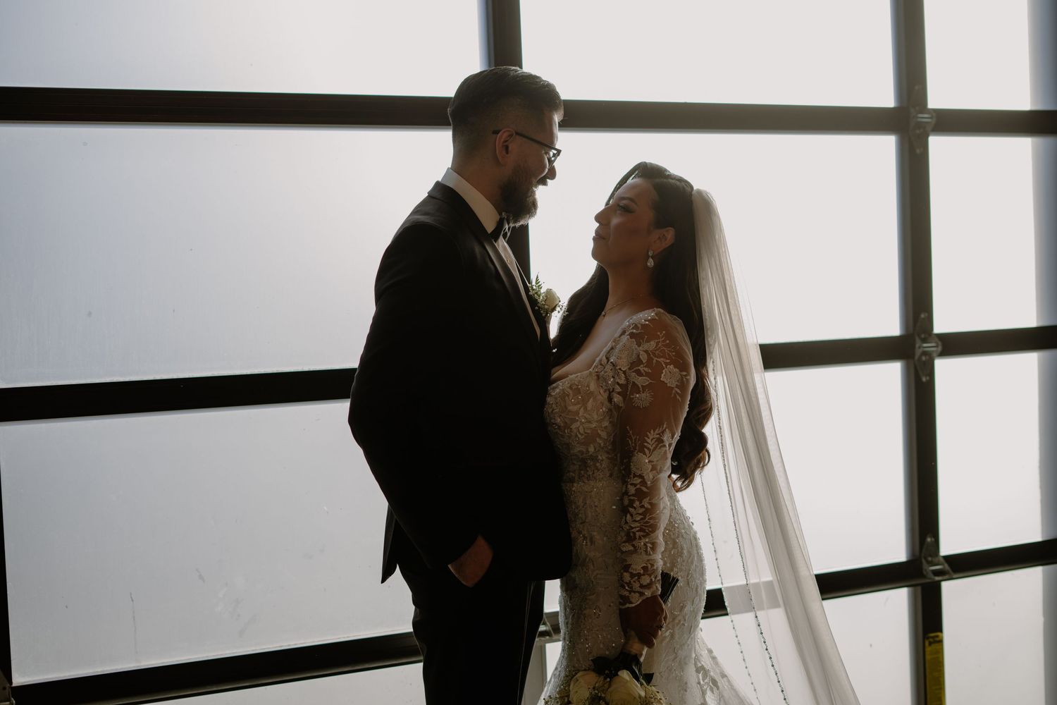 Wedding couple shares an intimate moment near large windows with natural backlighting.