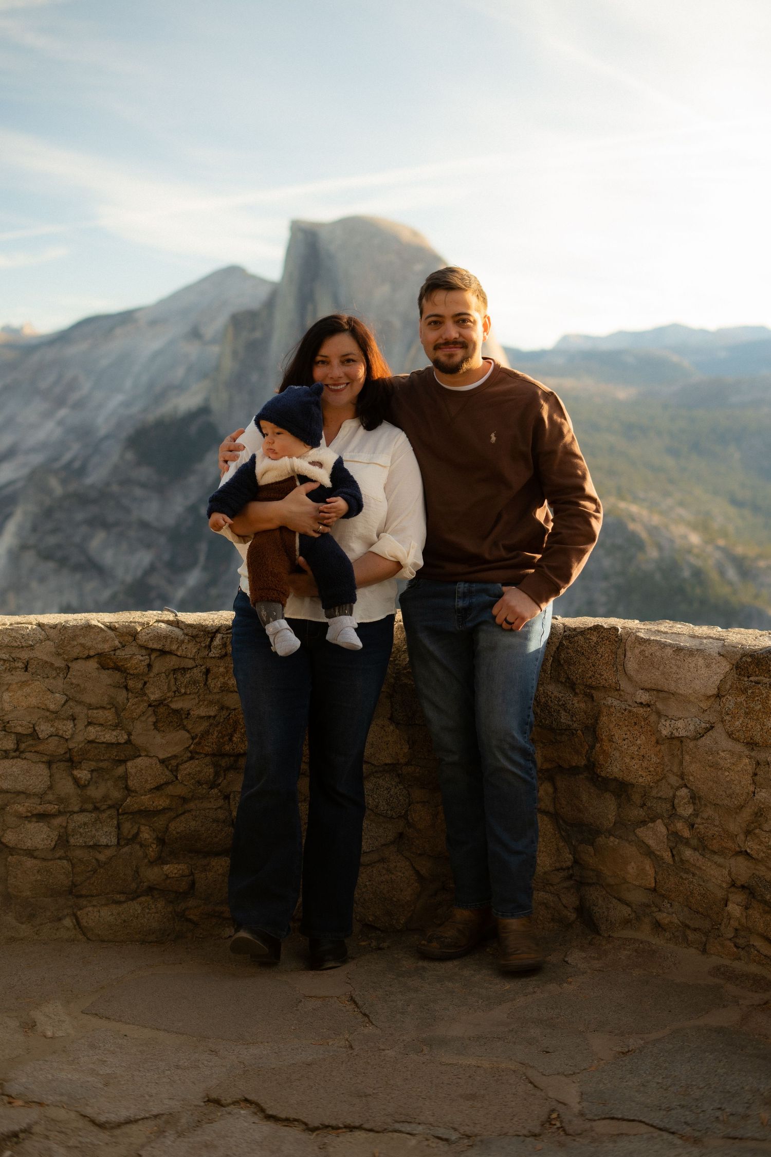 Family shares tender moment together against mountainous backdrop and stone wall.