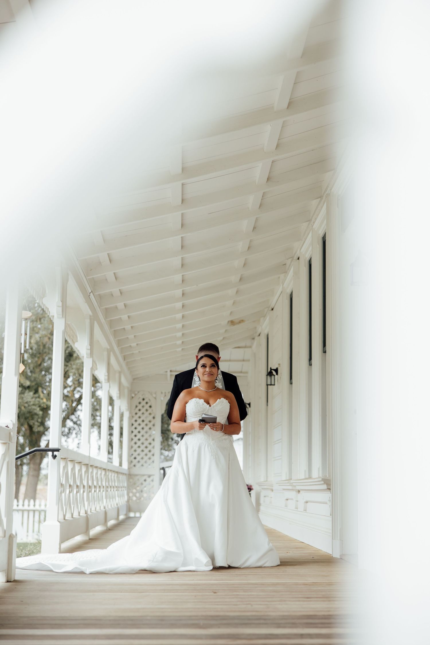 Bride and groom pose on a white colonial veranda, bride in elegant strapless gown with long train.