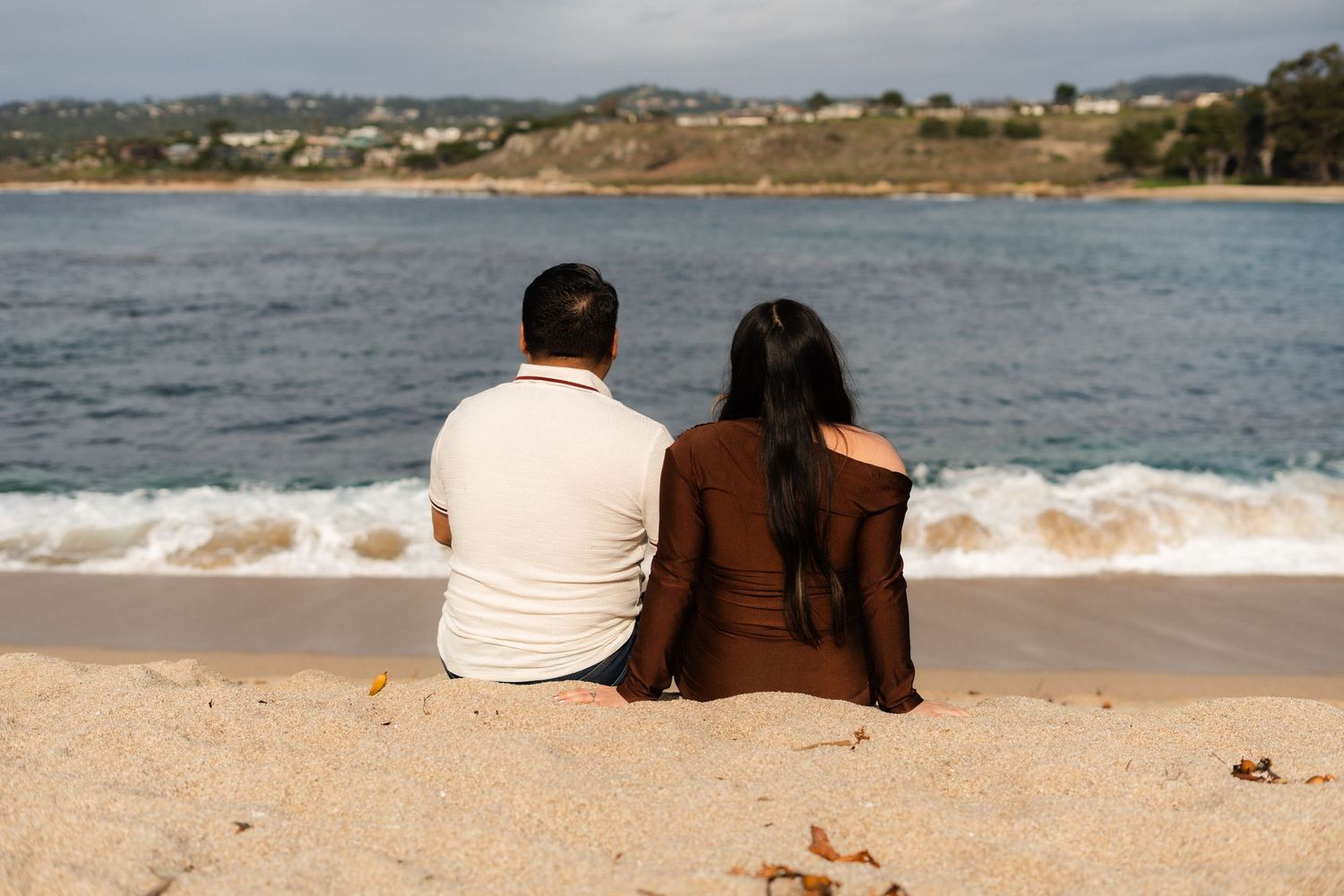 Two people sitting close together on a sandy beach overlooking the ocean.