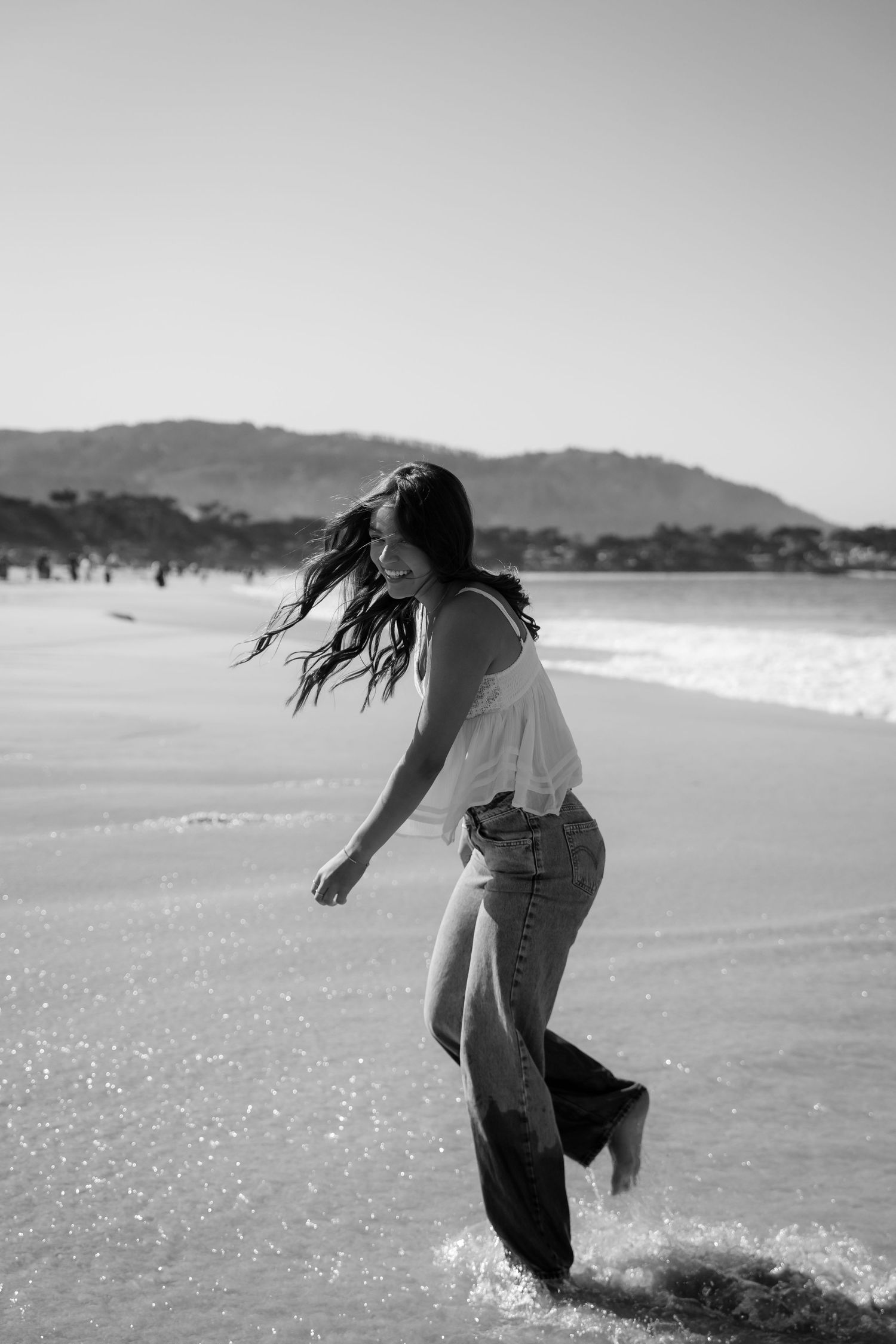 A carefree figure in jeans and white top splashes in the ocean waves at a scenic beach with mountains in the background.