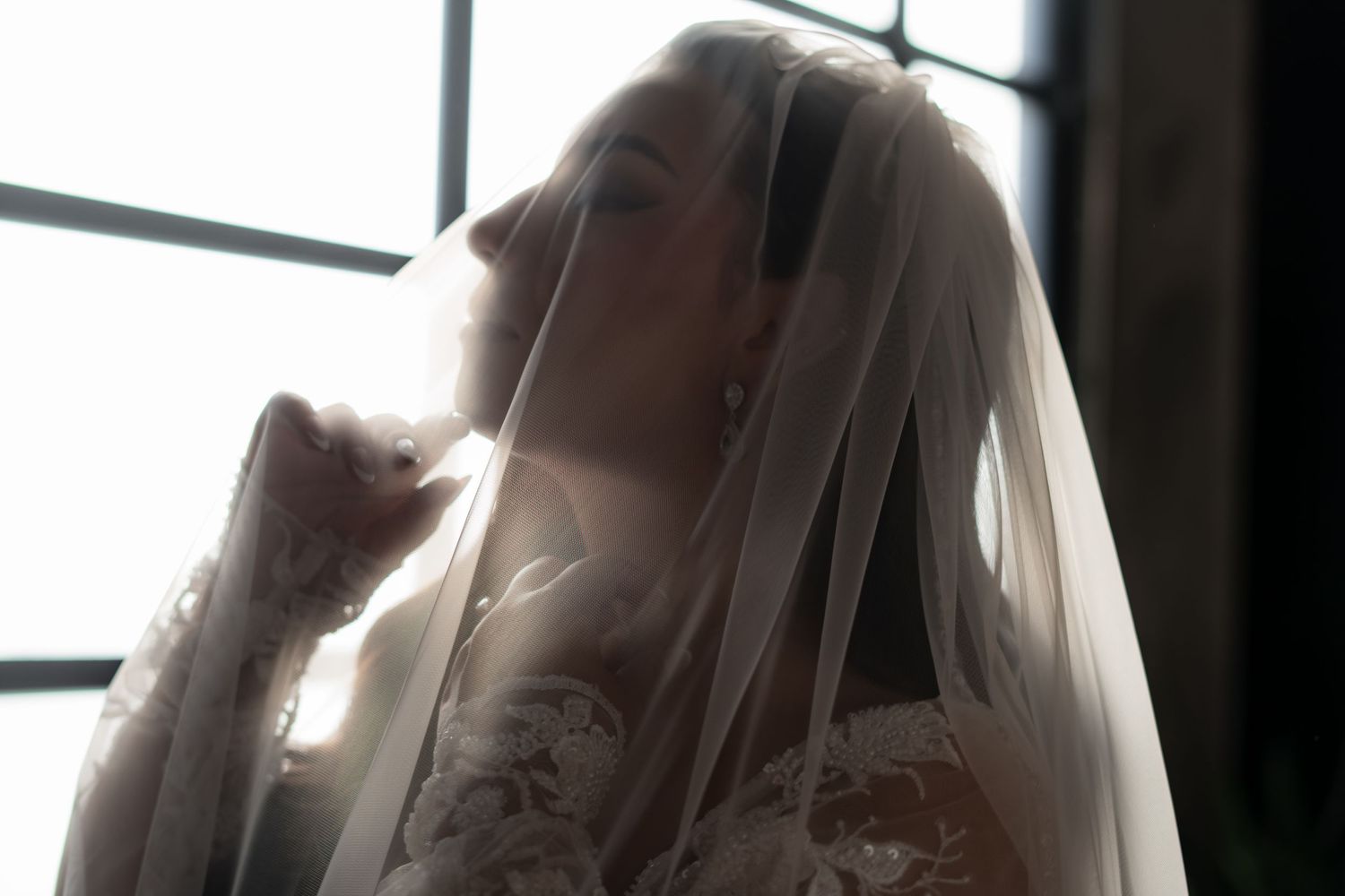 Dreamy side profile of a bride in her wedding gown with flowing veil against window light.