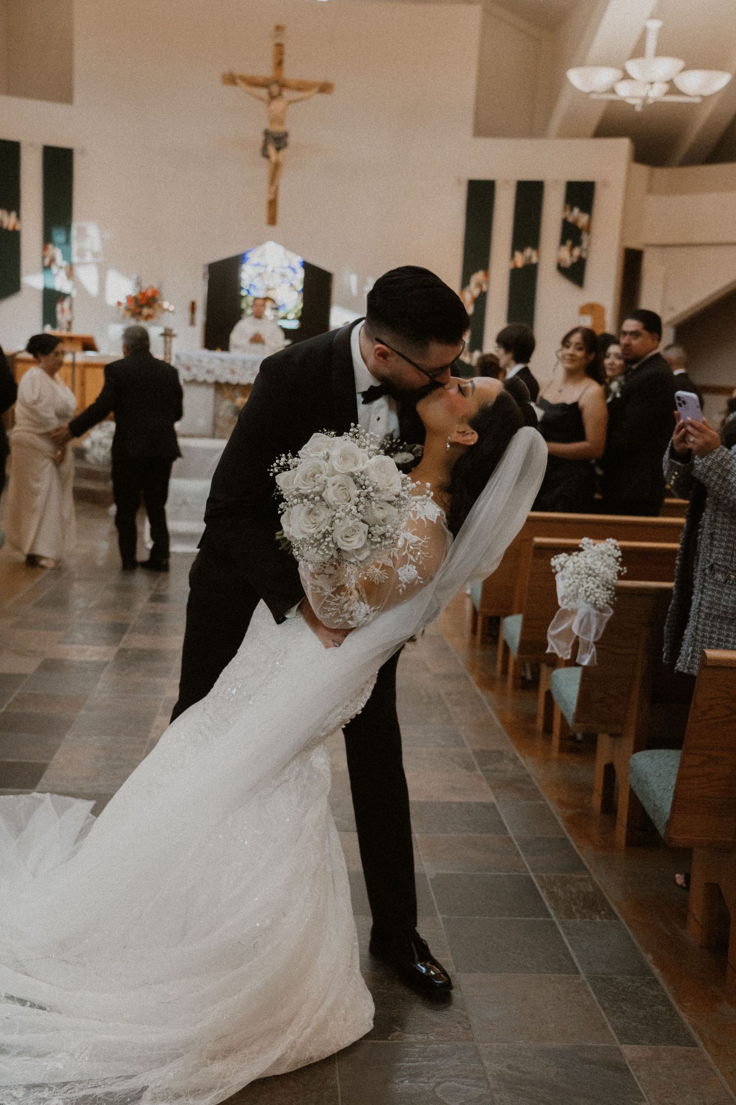 Bride in a lace dress and groom share a series of dips and kisses in a church aisle.