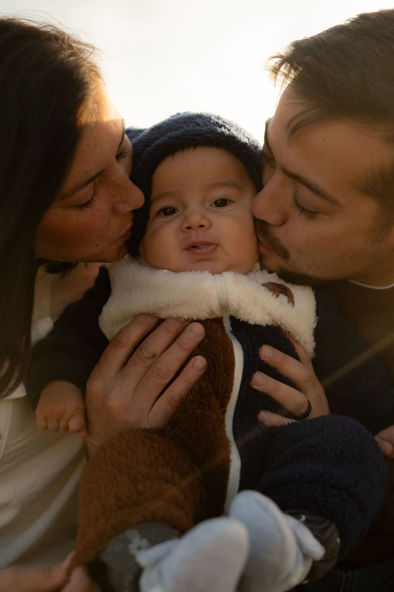 Sweet moment of parents kissing baby's cheeks in warm sunlight outdoors.