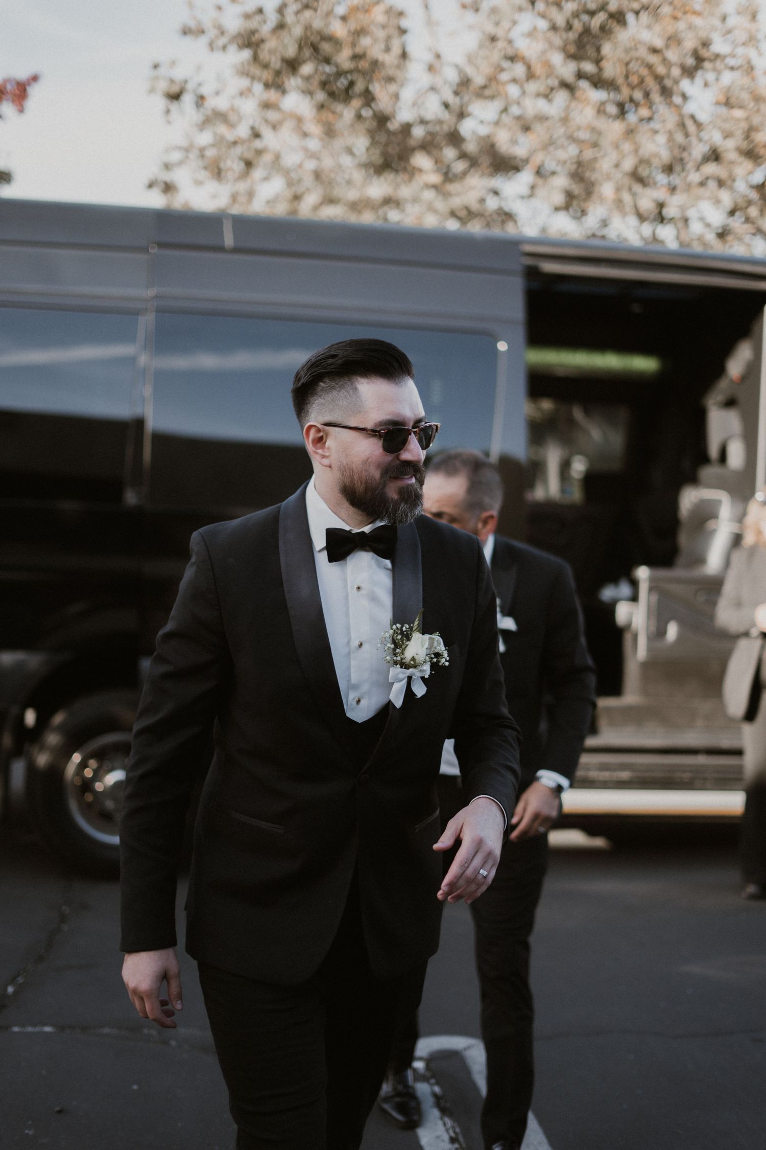 Formal wedding attire with black tuxedo, bow tie and boutonniere in outdoor setting.