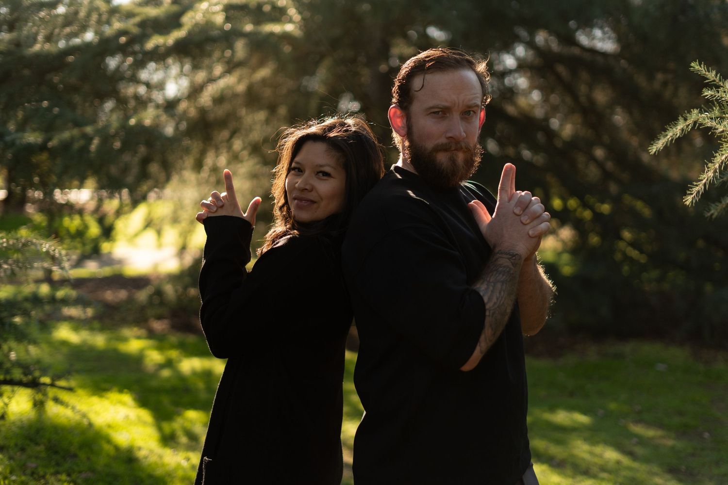 A playful couple posing back-to-back with finger guns in a park setting.