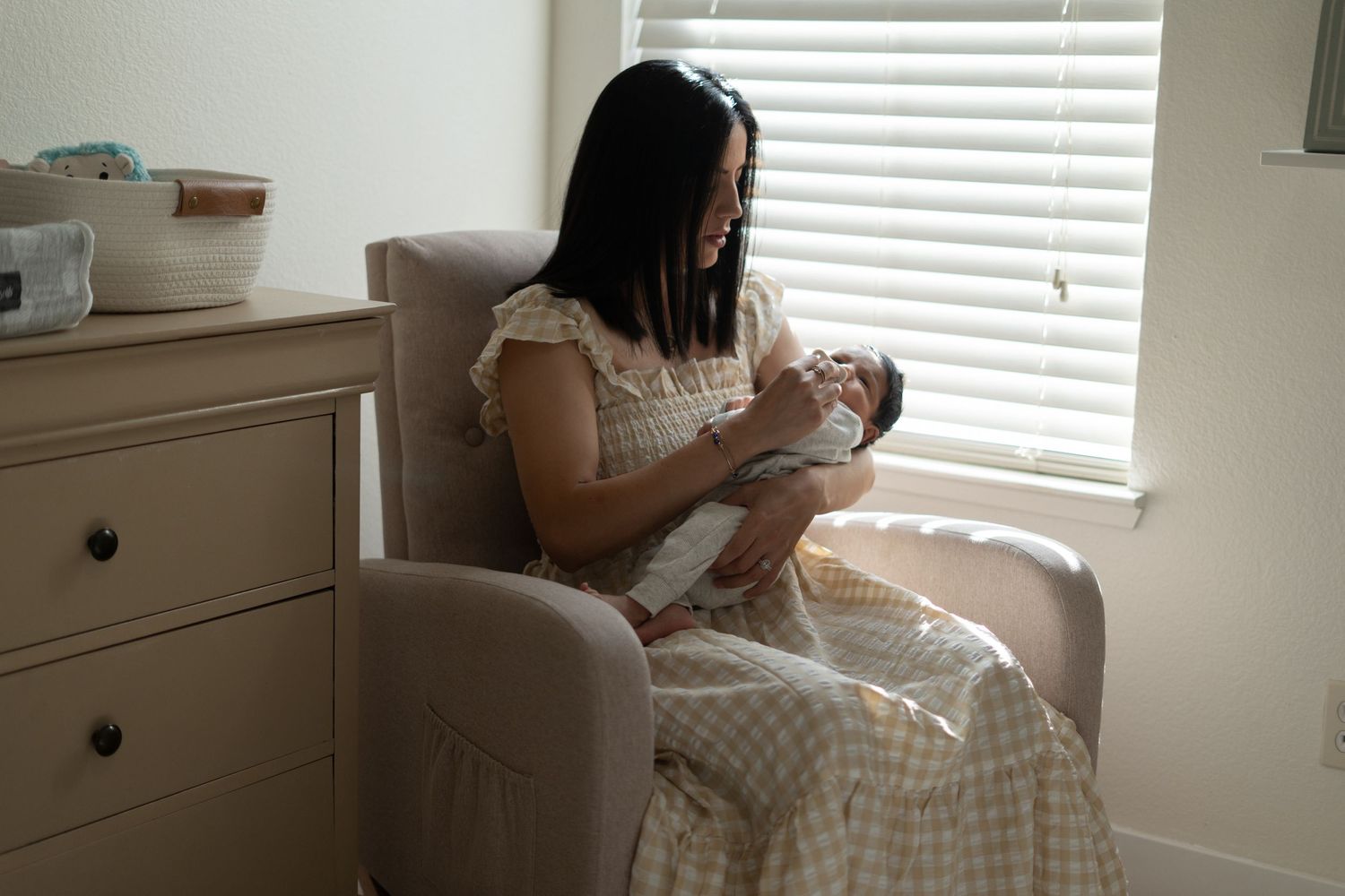 A mother in a gingham dress sits in a nursery rocking chair by a window while feeding a newborn baby.