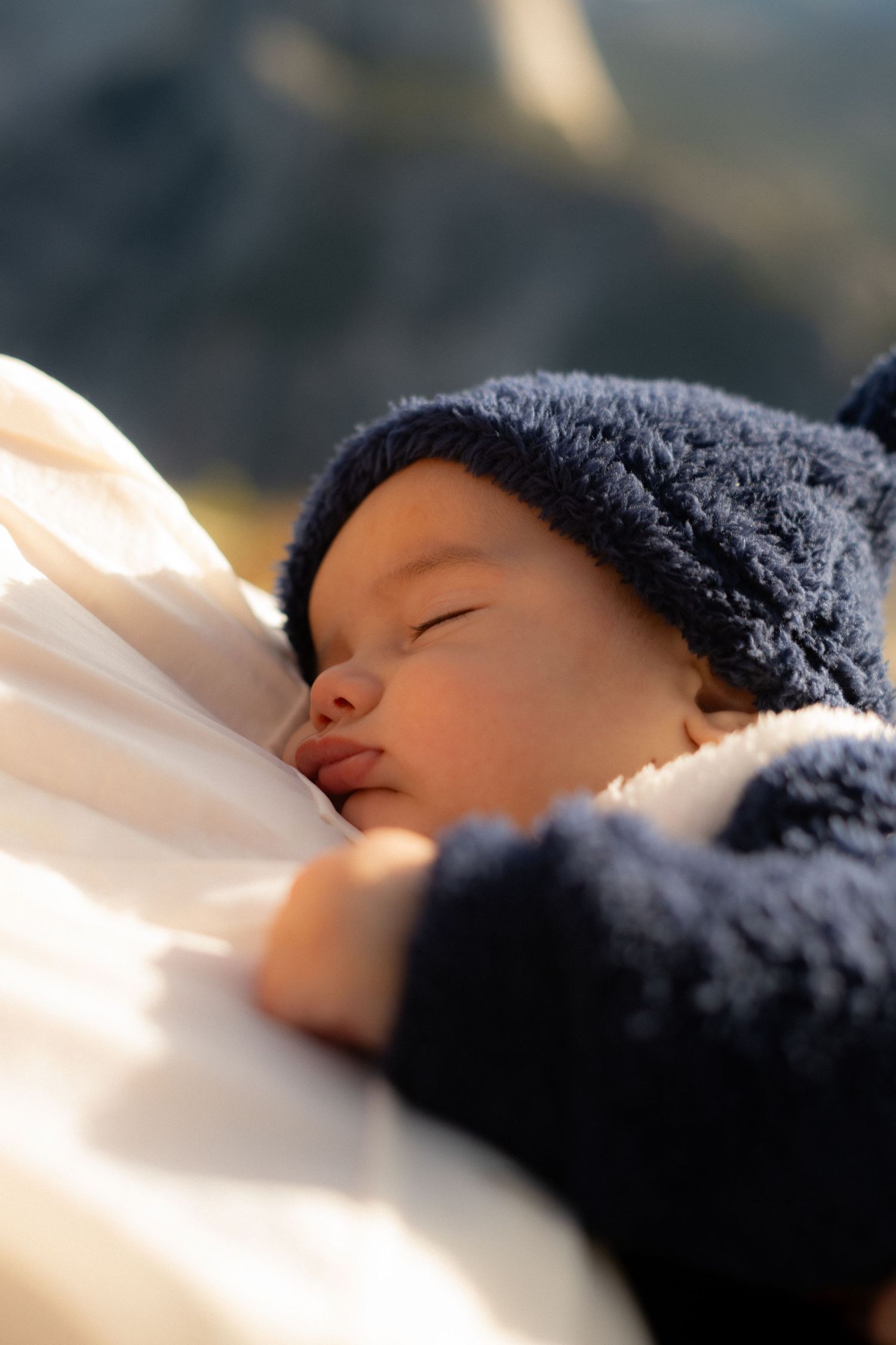 Baby wearing a navy knit hat peacefully sleeping against white fabric in warm sunlight.