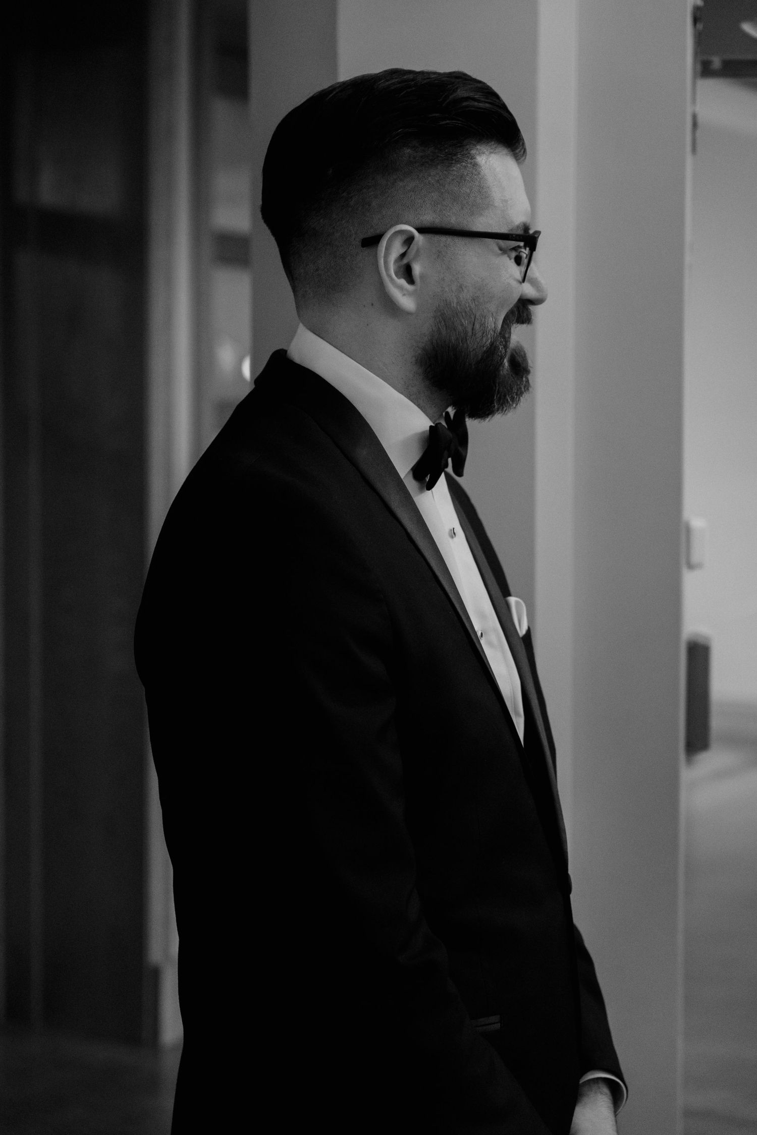 Close-up profile view of a groom in a black suit and glasses standing in a dimly lit hallway.