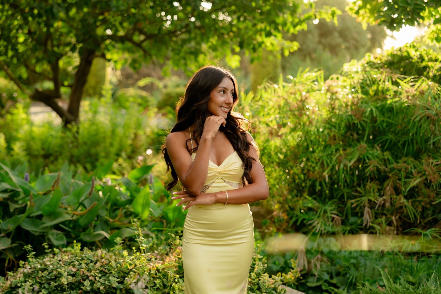 A person in a yellow dress poses in a lush garden setting during golden hour sunlight.