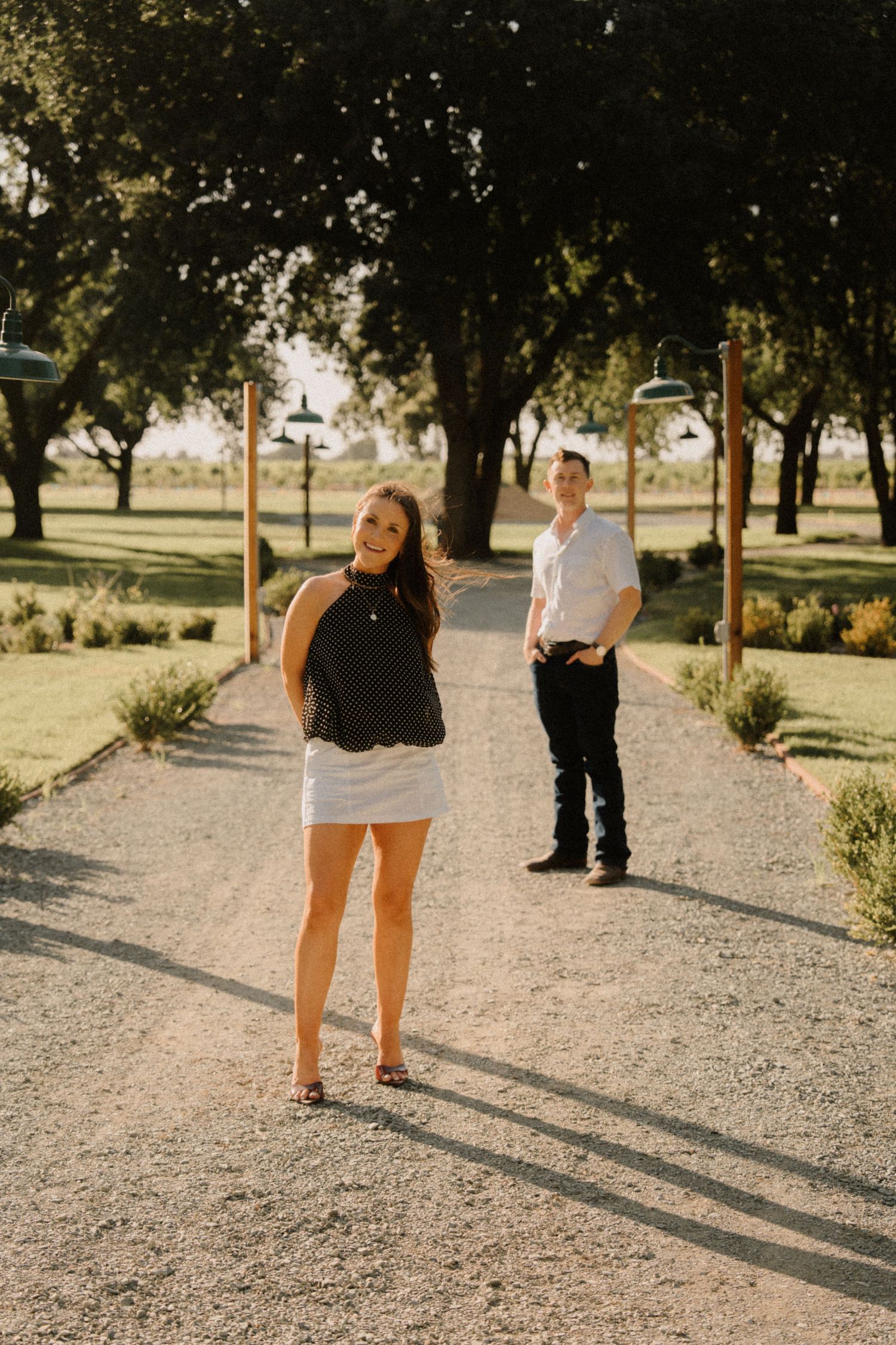 A smiling woman in a black top and white skirt stands on a gravel path, with a man in white shirt behind her.