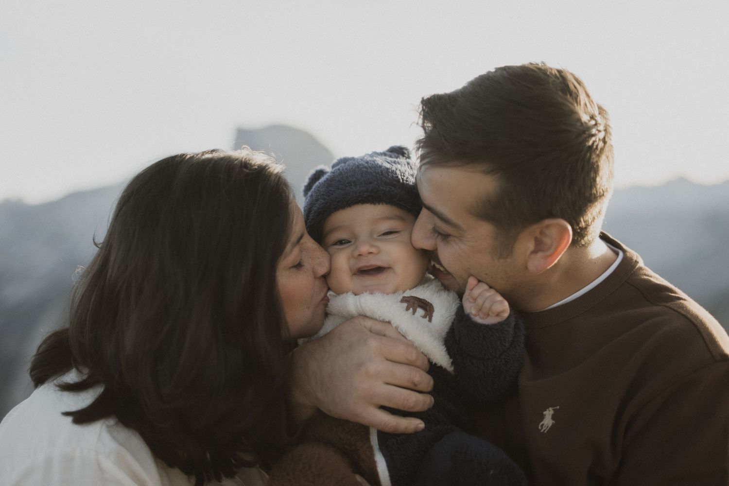 Family shares intimate moment with baby wearing blue hat against mountain landscape.