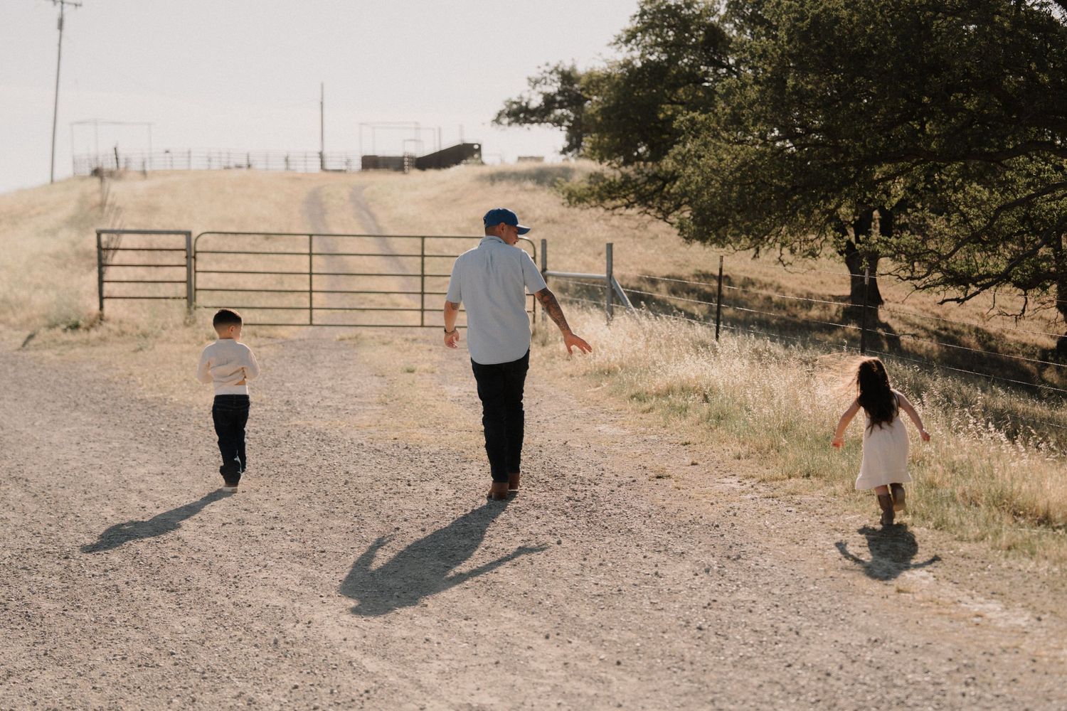 Adult and two children walking on a rural dirt path near a metal gate and oak tree on a sunny day.