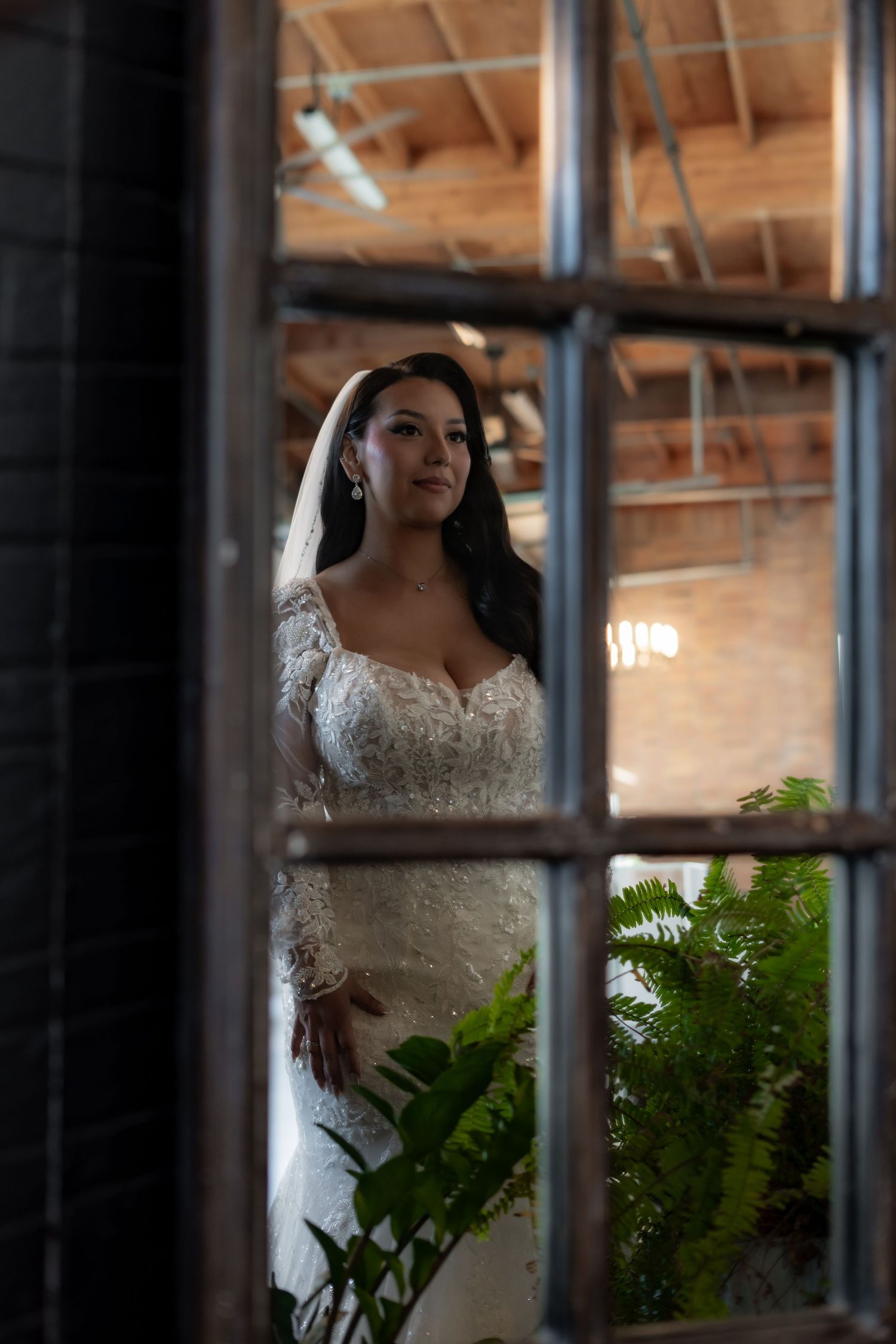Wedding dress detail seen through window panes with ferns and indoor lighting creating a romantic atmosphere.