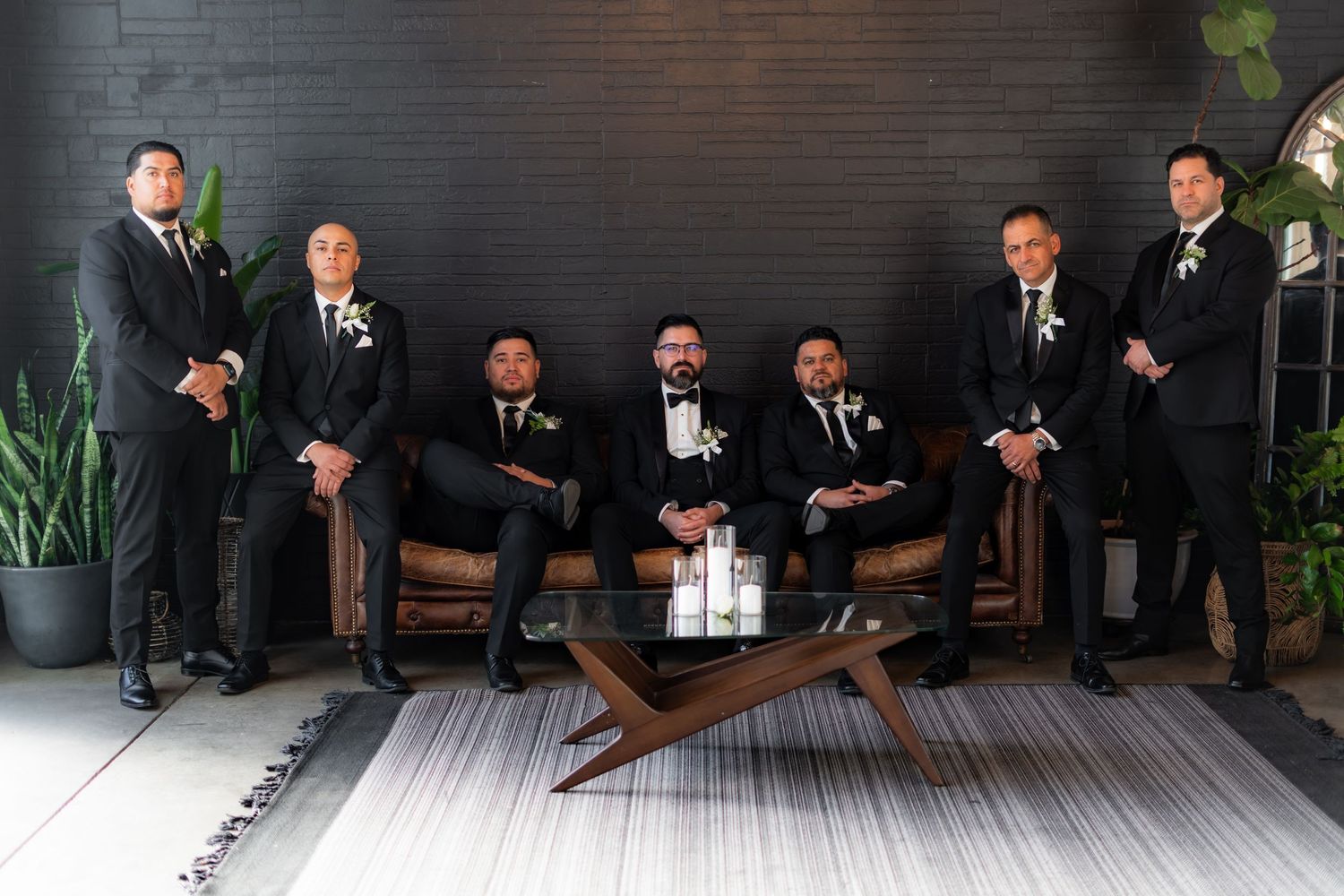 Formal group photo of wedding party in black suits arranged around modern coffee table.