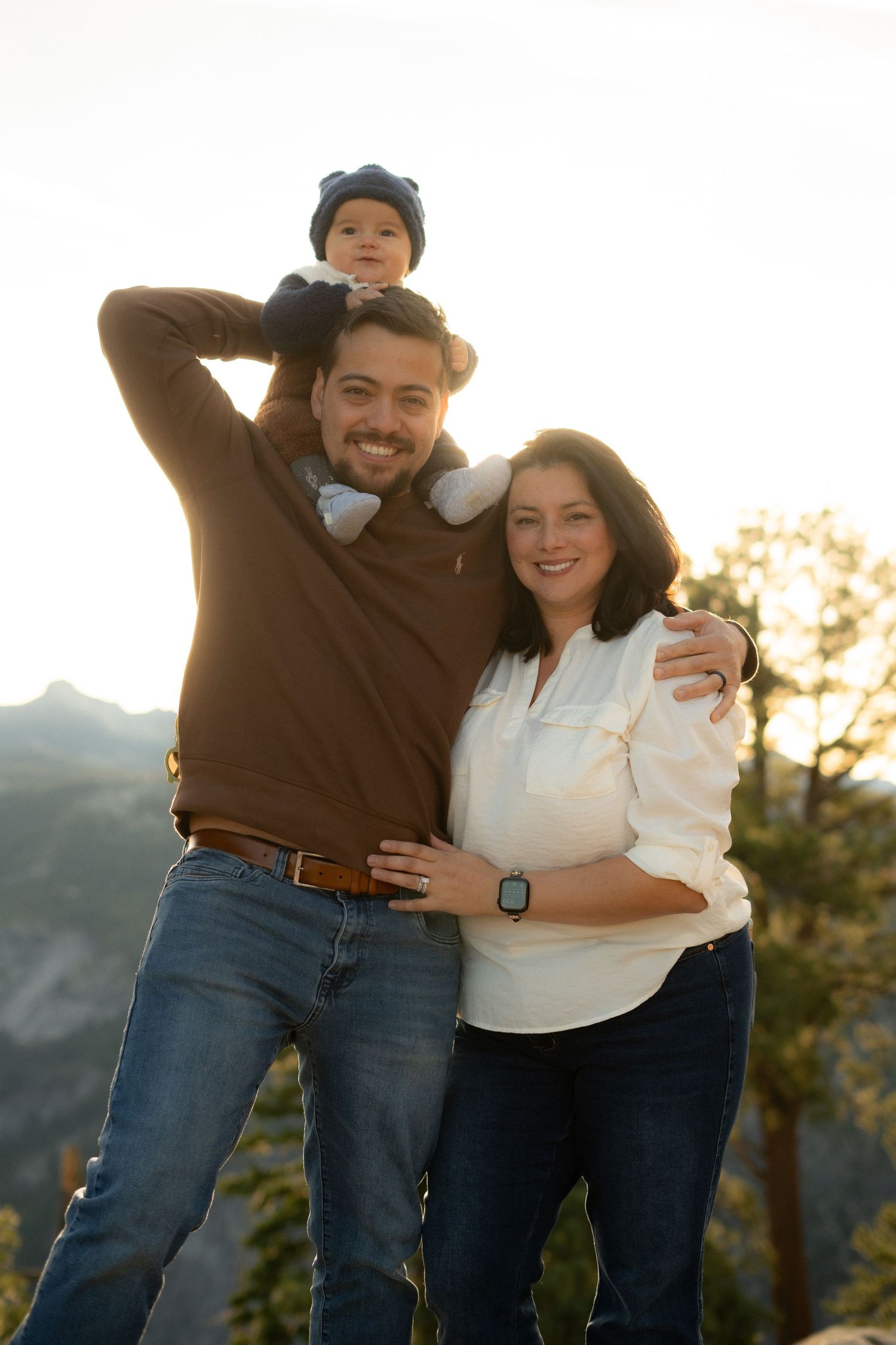 Joyful family portrait outdoors with baby sitting on shoulders against mountain landscape.