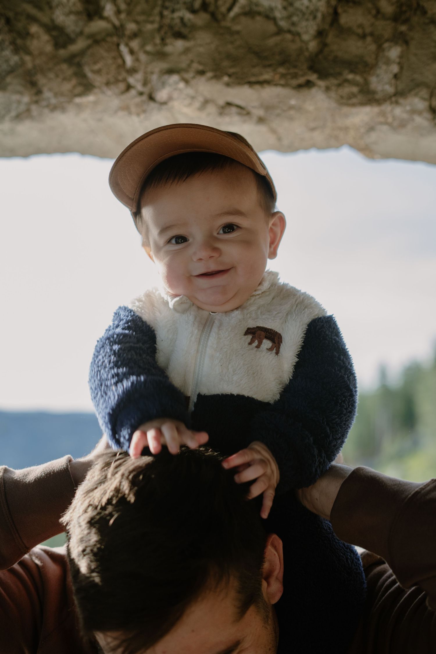 Baby wearing baseball cap and fleece jacket smiles while being held up.