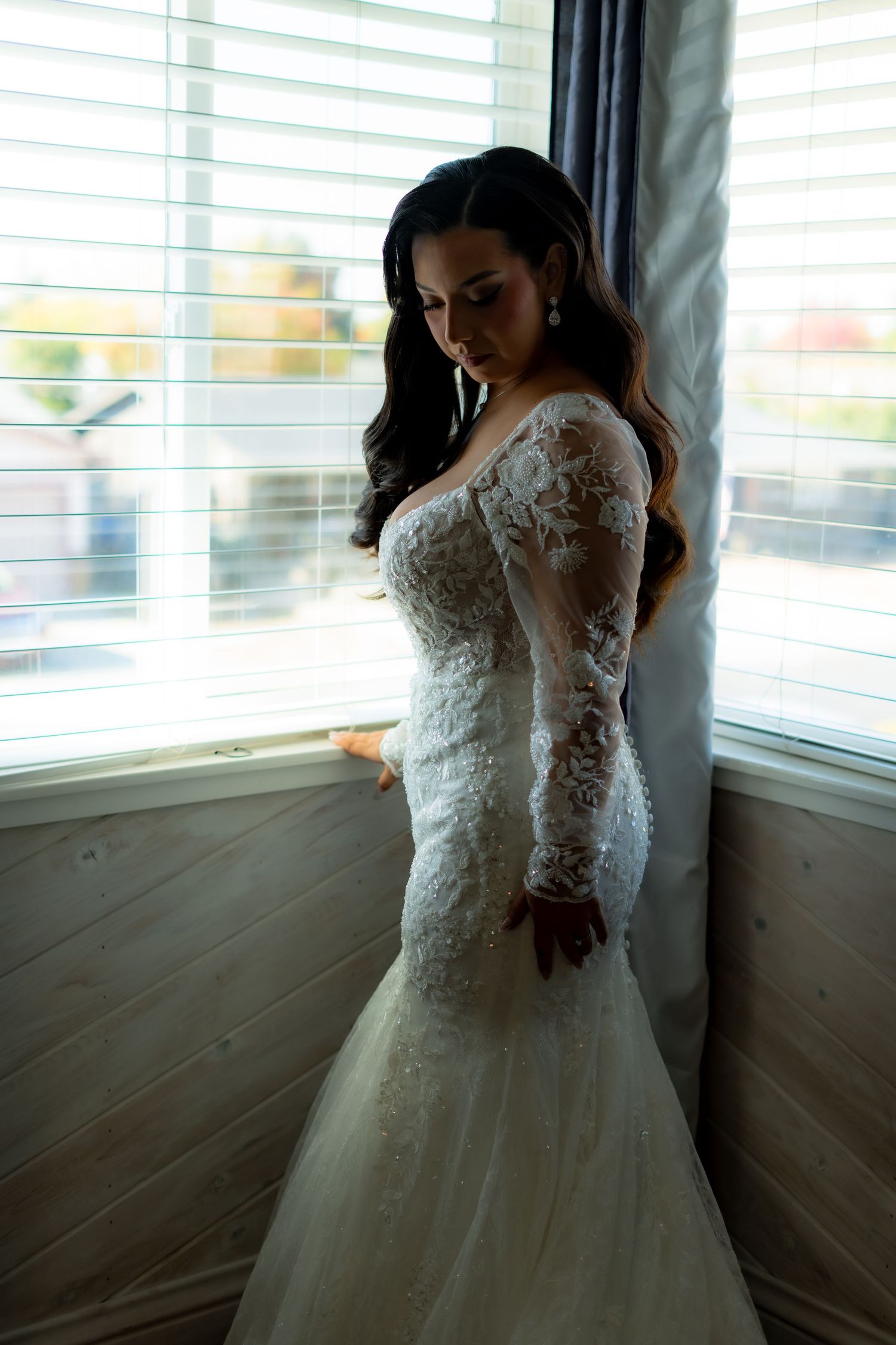 Bride in lace wedding gown poses near window with natural lighting.