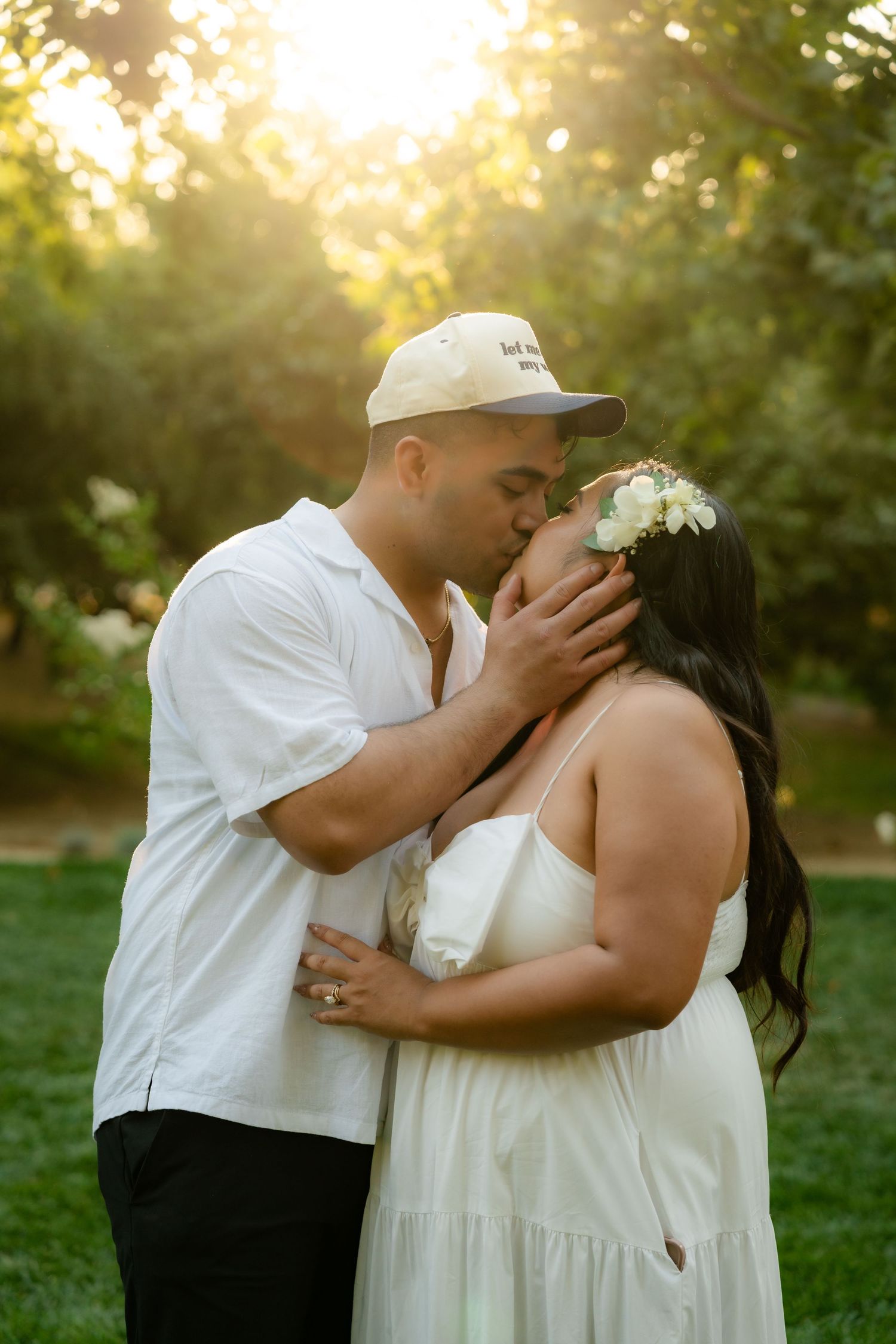 A romantic couple shares a tender kiss in a sunlit garden setting wearing white clothing during a special occasion.