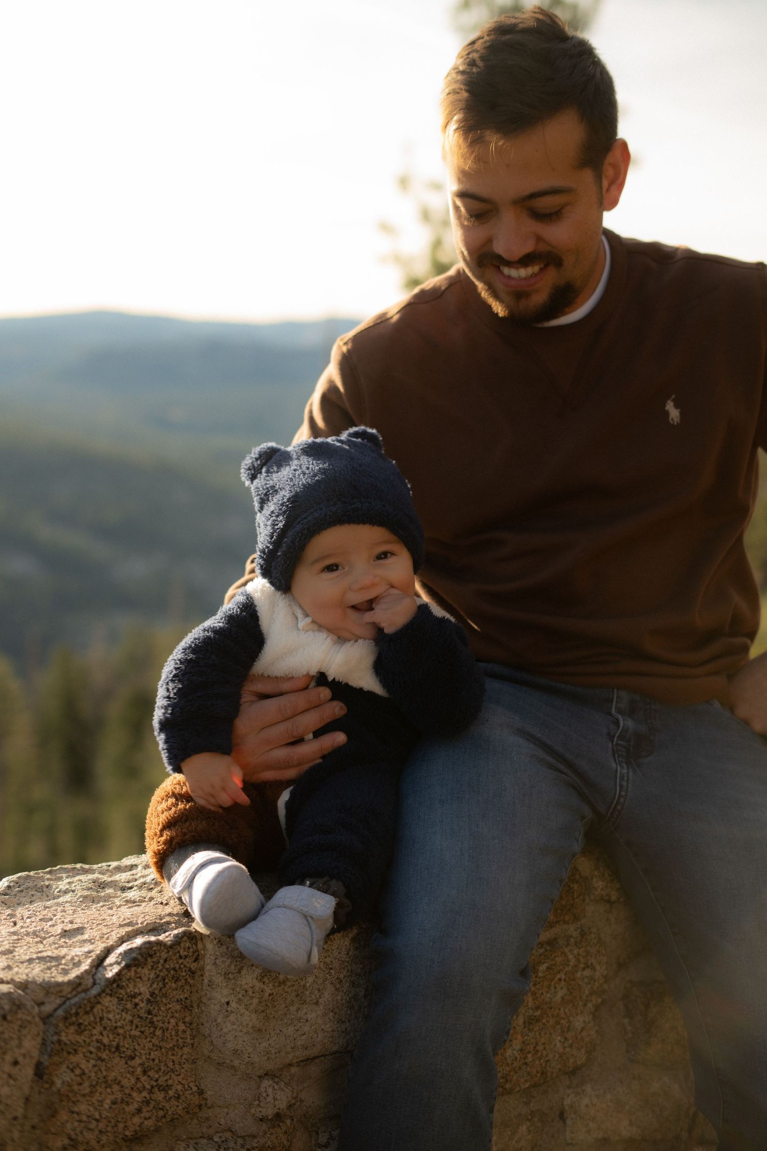 Candid moment between parent and baby sitting on rock during golden hour.