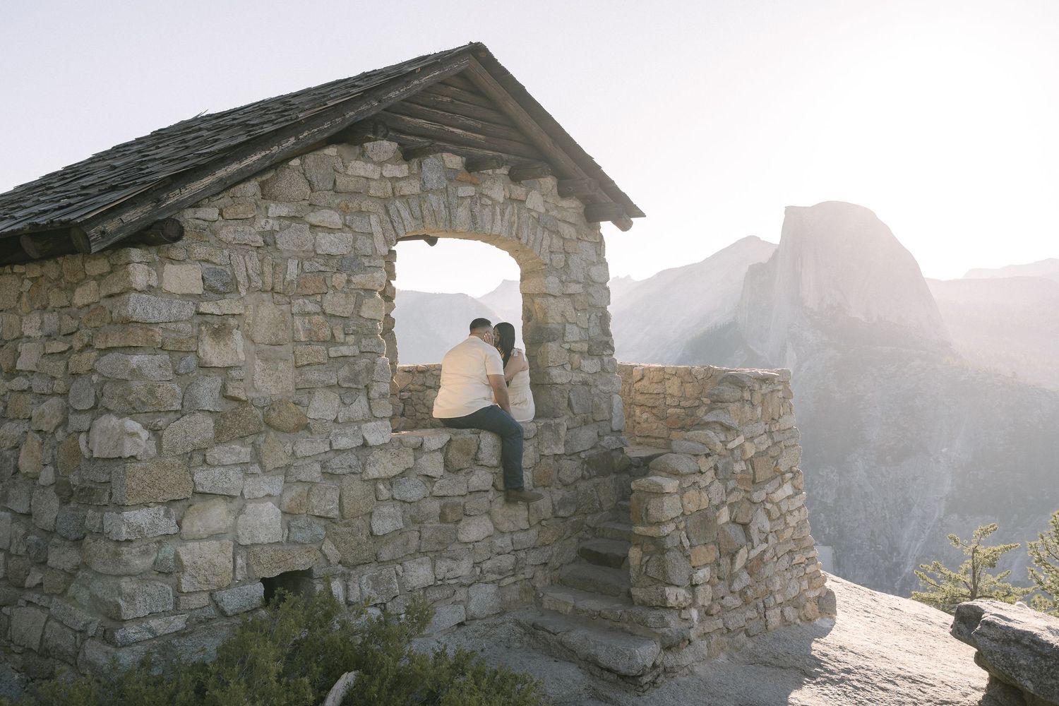 A scenic overlook from a rustic stone shelter at sunset shows misty mountain peaks in Glacier Point, Yosemite National Park.