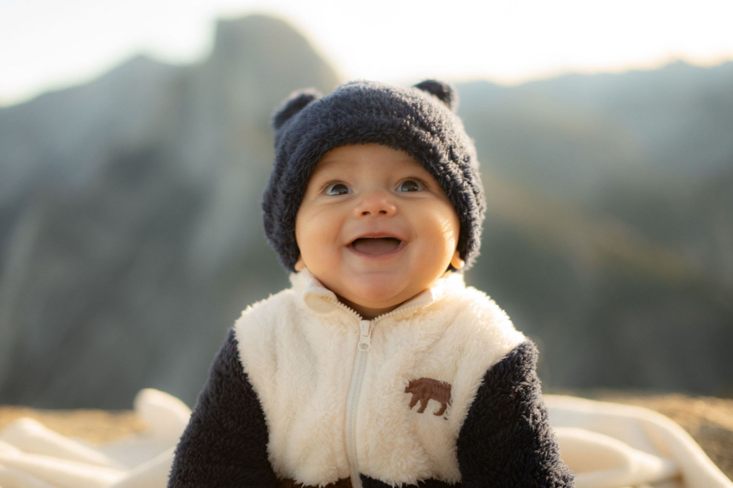 Smiling baby in navy beanie and bear-themed outfit against mountain backdrop.