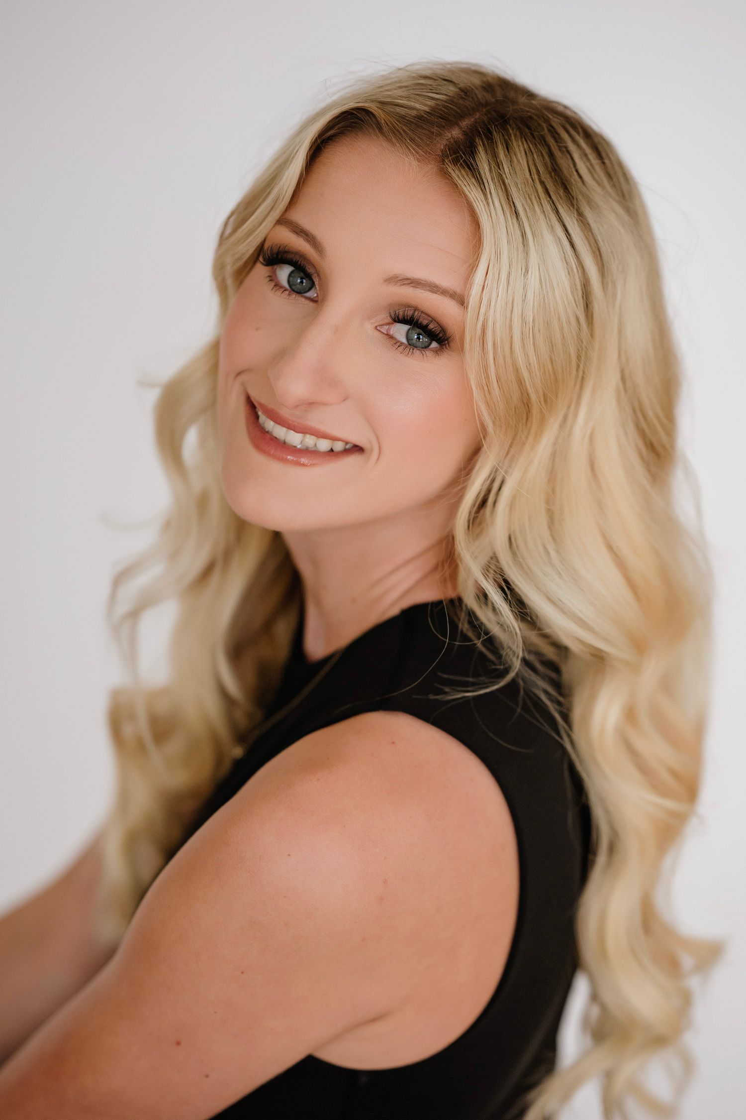 Smiling blonde woman with blue eyes and wavy hair wearing a black sleeveless top against a white background.