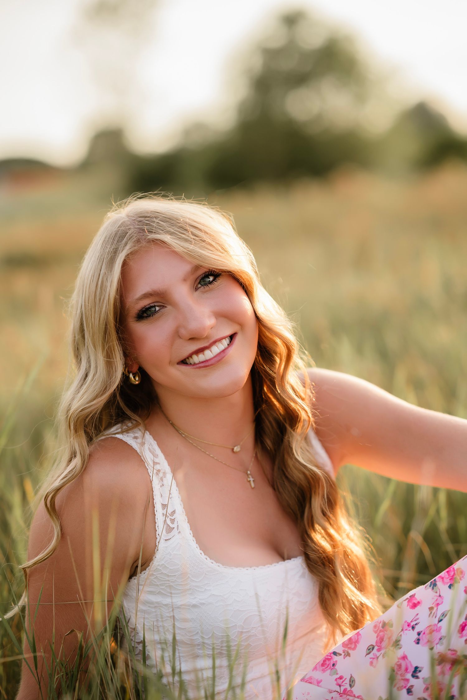 Smiling blonde teenage girl in white lace top sitting in golden field at sunset for senior portrait.