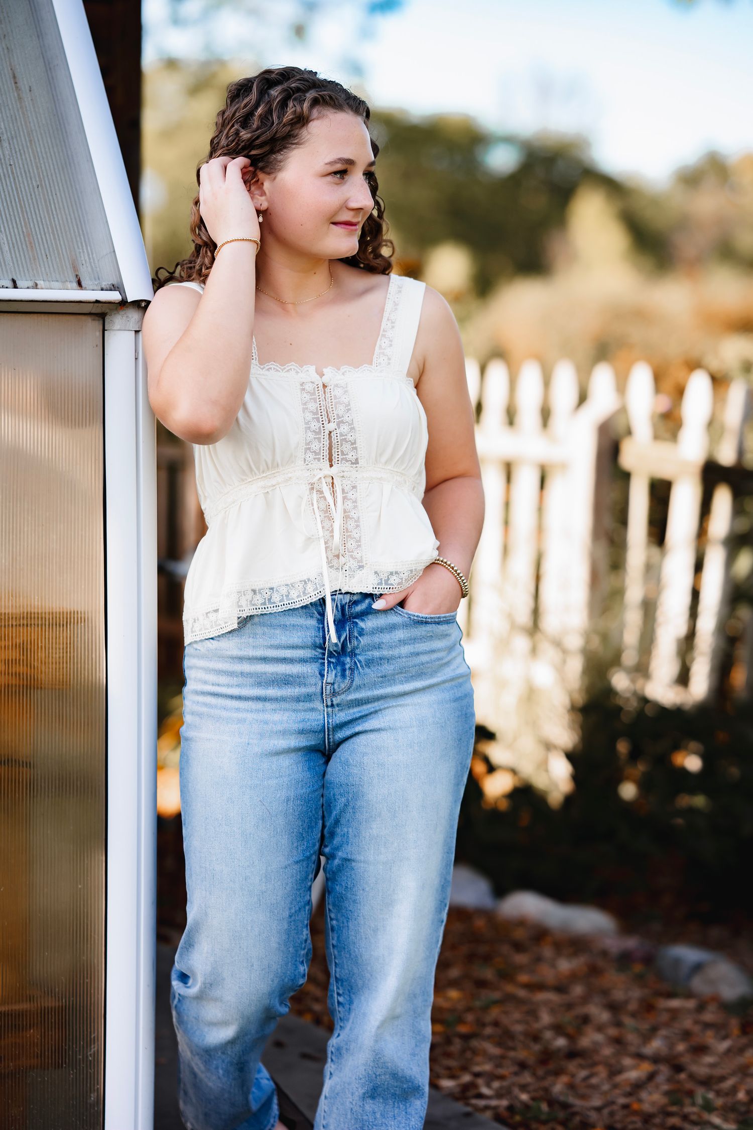A model in a white lace top and light wash jeans poses against a white picket fence during a casual outdoor photoshoot.