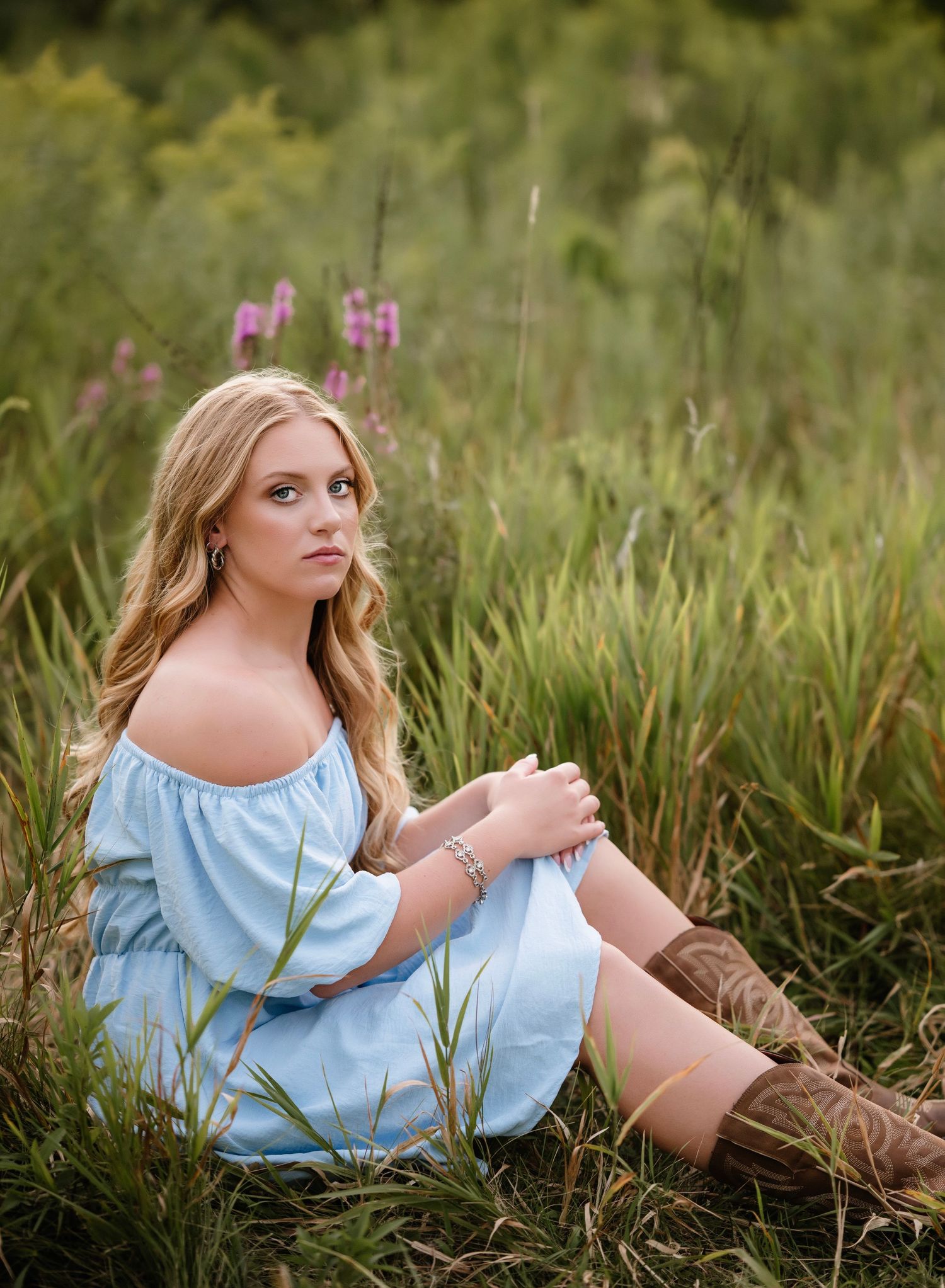 A person in a light blue off-shoulder dress sits gracefully in tall grass with wildflowers during a summer photoshoot.