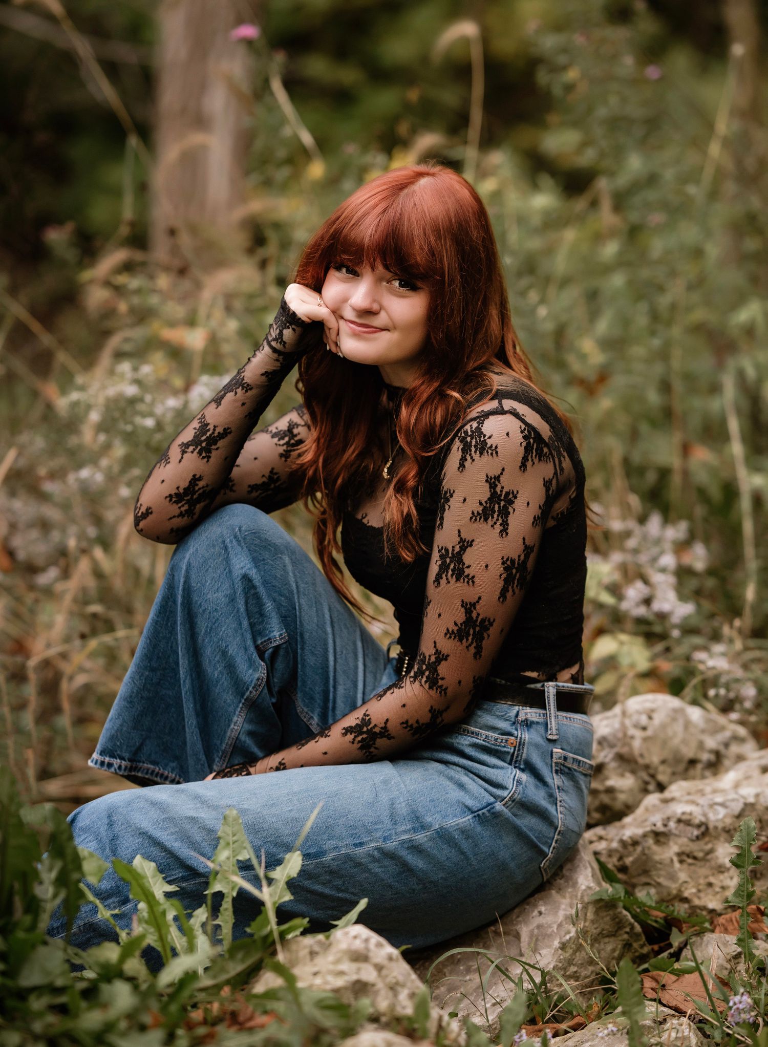 A person in a black lace top and jeans sits outdoors among wildflowers and greenery on a sunny day.