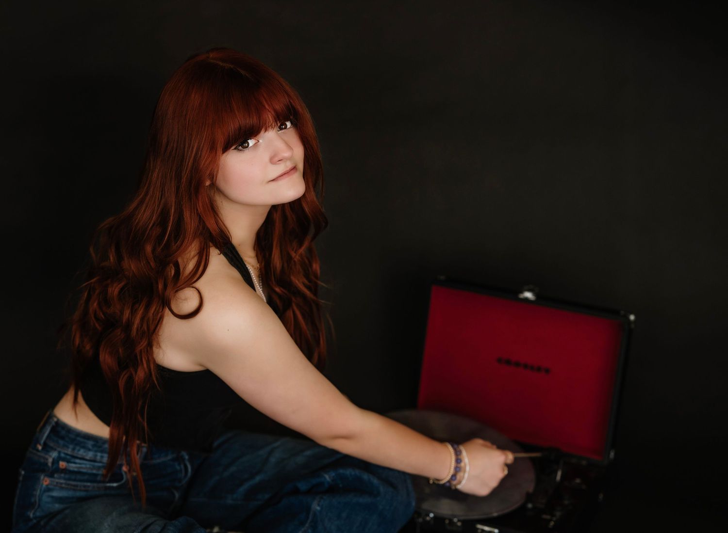A person with long red hair poses against a dark background while holding a burgundy colored laptop.